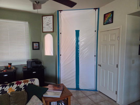 A portion of a formal living and desk area with green walls. The open space and wall cutout window to a foyer are sealed off with white plastic sheeting with a blue zipper line.