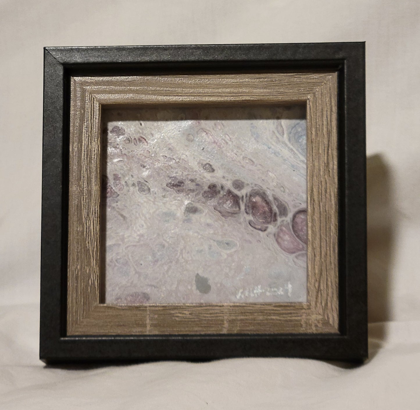 A picture frame with black outer framing and brown wood-grain inner framing. The featured picture is a fluid art square featuring deep purple bubble in a largely lavender stretch. There are small light blue bubble in the lavender and dark gray threading around many bubbles. In silver in the lower right corner are the artist's initials "KMH" and the year completed "2024."