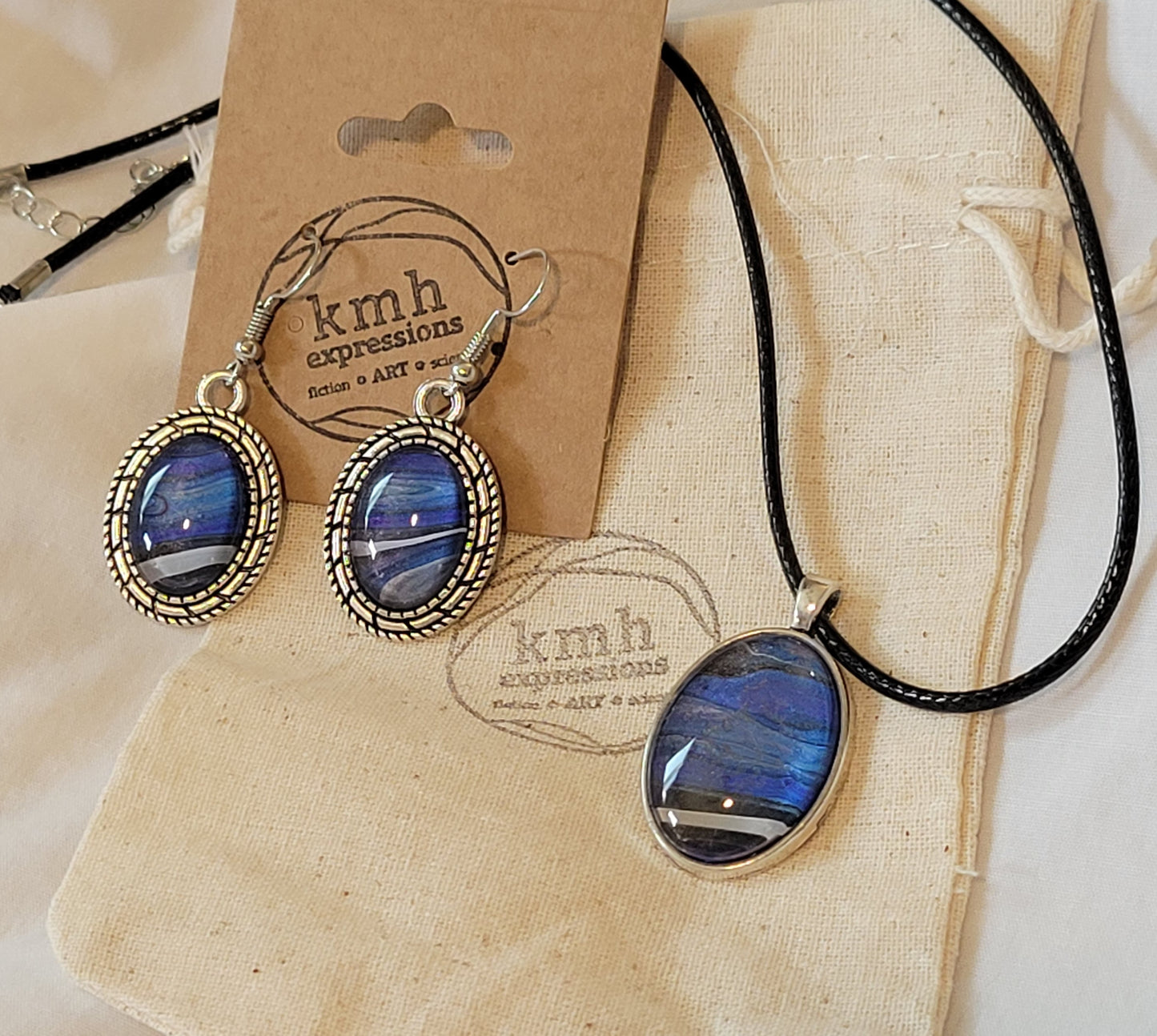 A jewelry set of a necklace and earrings. The necklace is an oval cabochon stainless steel pendant with a black faux leather cord; the coloring of the pendant is striations of blues and purples streaking from one side to the other with stark bands of black and white at the base. The earrings are oval cabochon stainless steel dangles with a segmented pattern border; the earrings have similar coloring to the pendant, only the streak of black and white stretches through them at an angle in alignment with each.