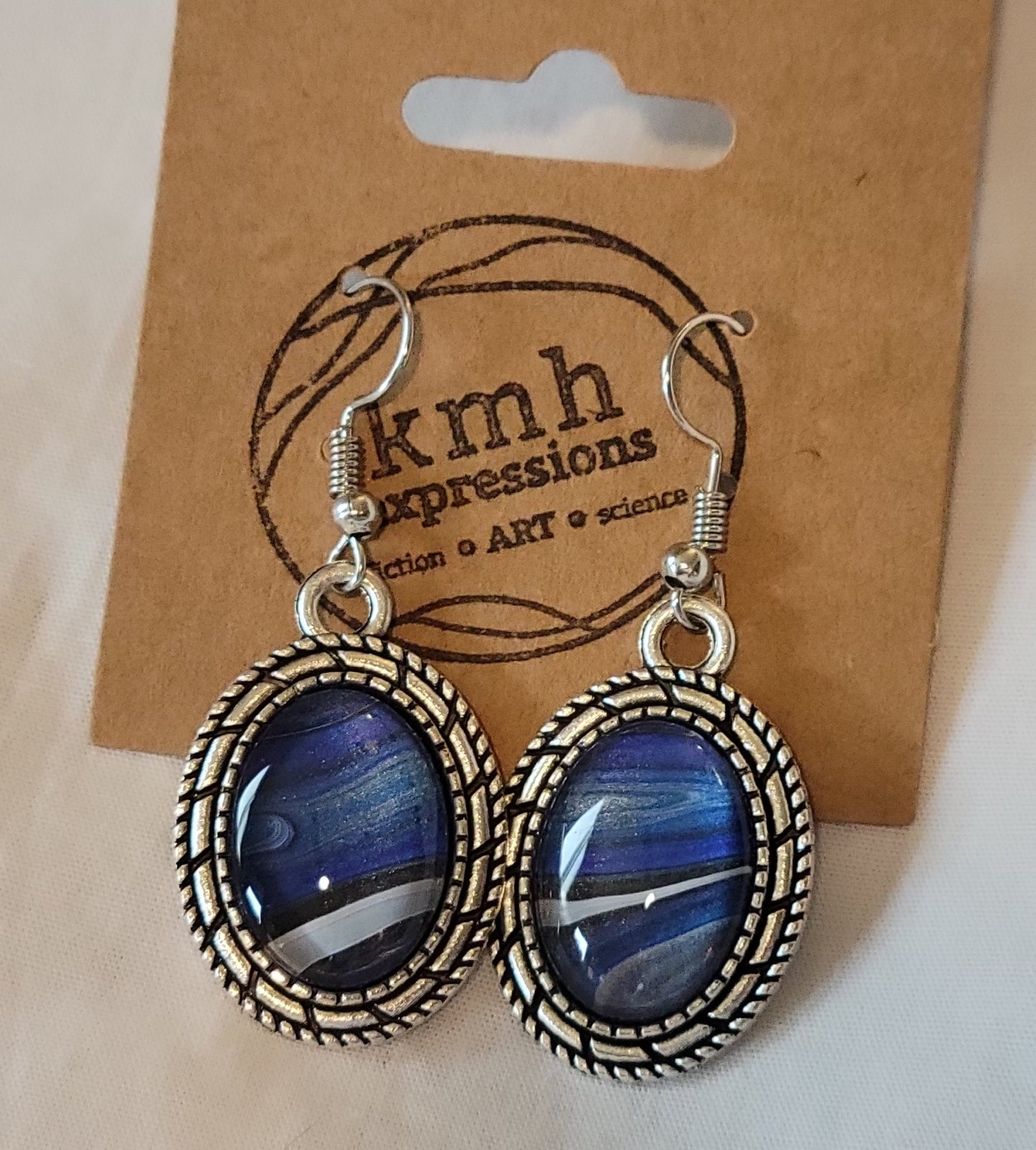 The oval pierced earrings dangle from a card stamped with the KMH Expressions logo. The earring posts are sterling silver and the base are stainless steel. They have a rope-like design around the cabochon. The earrings are aligned so the black and silver/white of the event horizon line up well against the hazy blue of the nebula clouds.