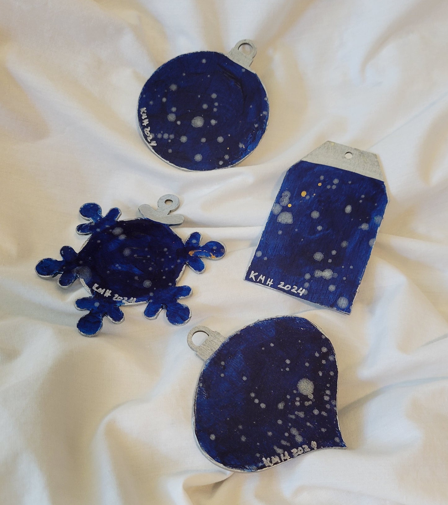 The reverse of the ornaments featuring dark blue background with silver speckles. Each ornament has the artist's initials "KMH" and year completed "2024" in silver toward the base.