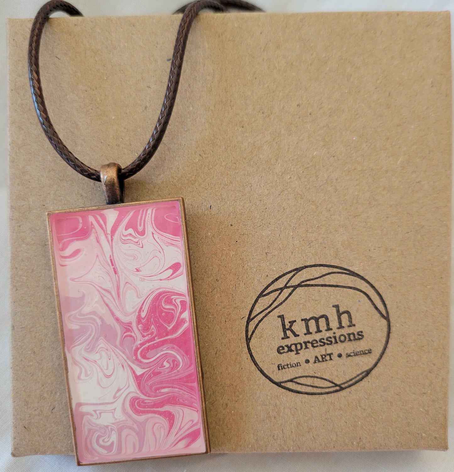 Rectangular pendant with a bronze setting and a brown imitation leather cord rest against a brown cardboard jewelry box stamped with the KMH Expressions logo. The pendant had swirls of dark pink, light pink, lavender, and white and is sealed with resin.