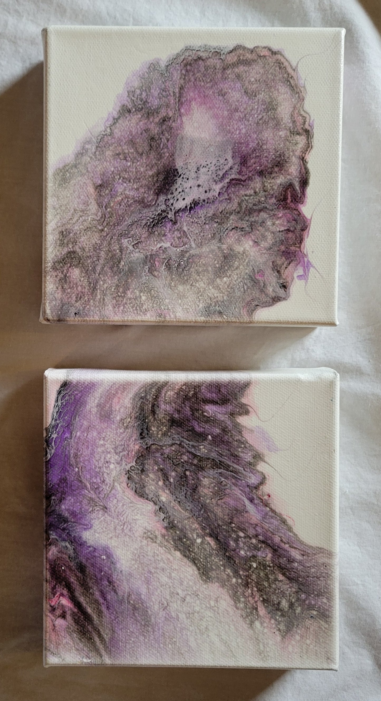 Two 6x6x1.5 in gallery wrapped canvases, both featuring fluid acrylic art in pinks, purples, grays, and blacks. The layers of color swirl and layer in a way that resembles a nebular, particularly when the two canvases are oriented together, as if the fluid art poured from one canvas to the next without interruption.