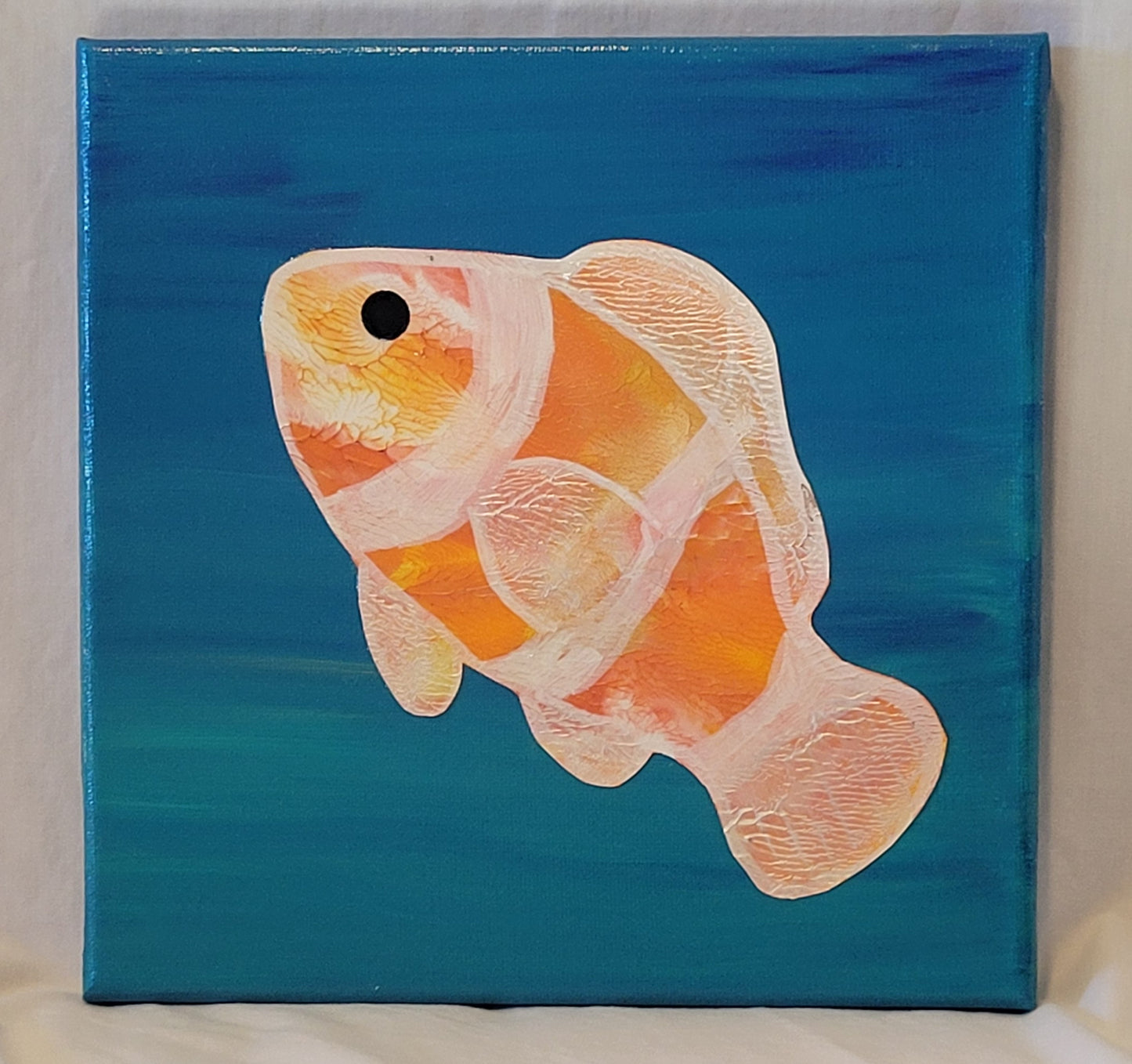 Canvas background painted with a blend of blues and turquoise that create a streaky lagoon look. A cutout of a clownfish is centered on the canvas. The cutout has been painted in oranges, golds, silvers, and pearls in a scaly pattern. A black alkyd oil dot creates an eye.