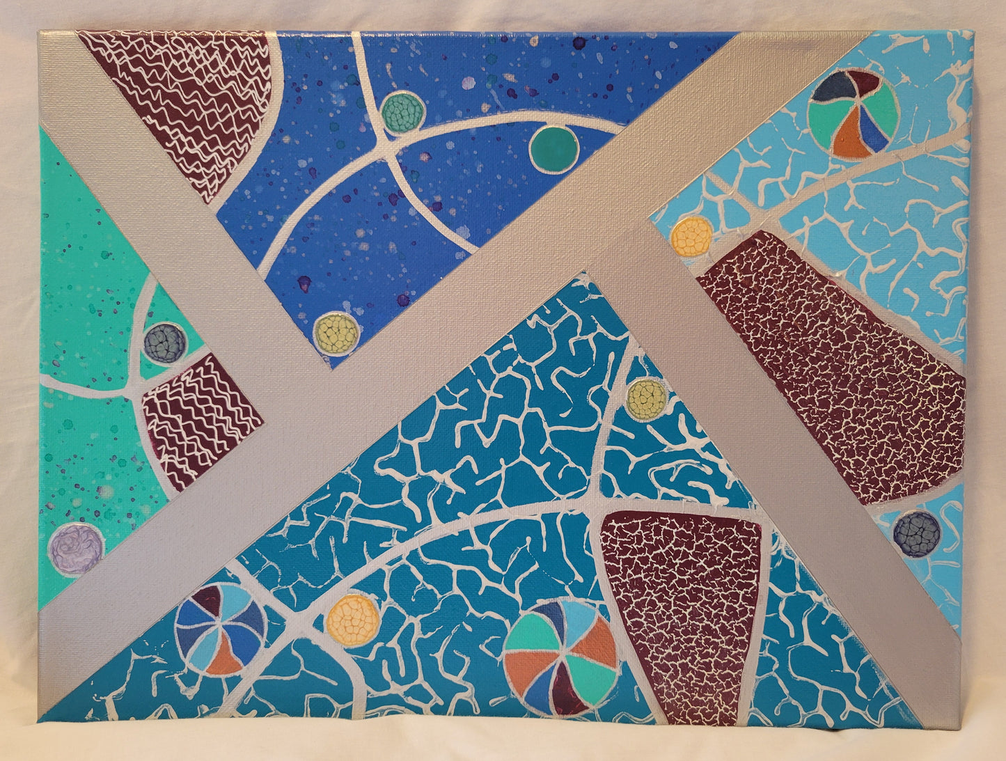Canvas split nearly diagonally with a silver band. There are two additional silver bands in a split diagonal that meet the central silver band. Each area created by the bands is painted a different shade of blue: aqua, turquoise, sky blue, deep blue. The aqua and deep blue sections are speckled with purples and silvers and greens. The turquoise and sky blue sections are stamped in white with a neural fractal pattern. Smaller areas are painted violet and covered in holographic foil or white wavy lines.