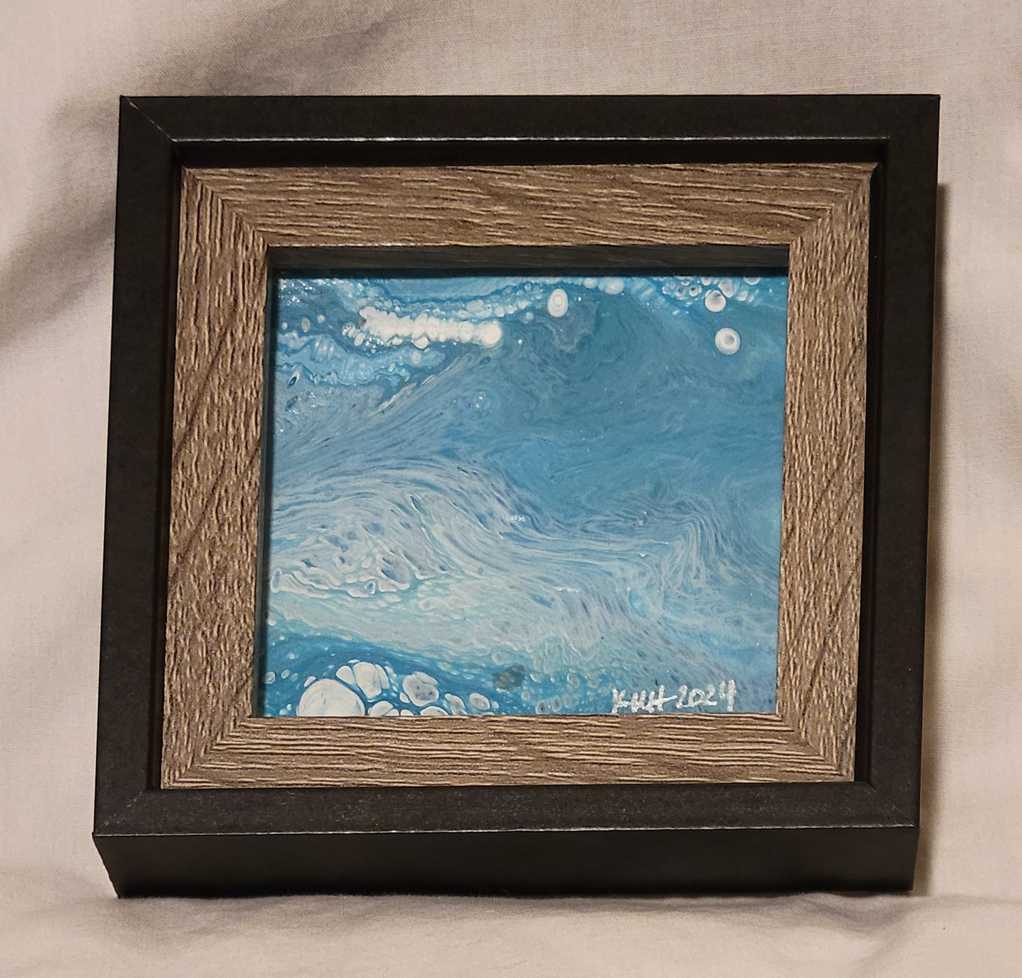 A picture frame with black outer framing and brown wood-grain inner framing. The featured picture is a fluid art square featuring blue shades with white and silver swirls and bubbles that give a sense of a wave crashing. In silver in the lower right corner are the artist's initials "KMH" and the year completed "2024."