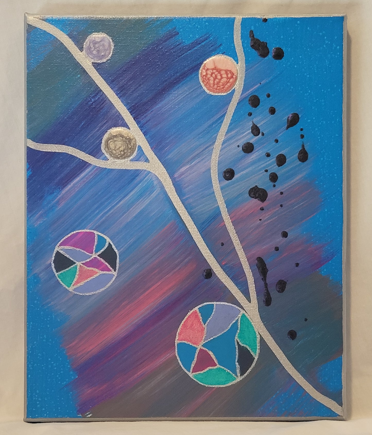 A canvas is painted a distressed bright sky blue with swipes of gray, purple, pink, and lavender fanning out from a silver diagonal line across the canvas. There are two silver branches reaching out to opposite edges of the canvas. Multi-colored balls and alkyd circles bud out from the branches and float away. Dried acrylic drips in dark purple dot on side of the canvas.
