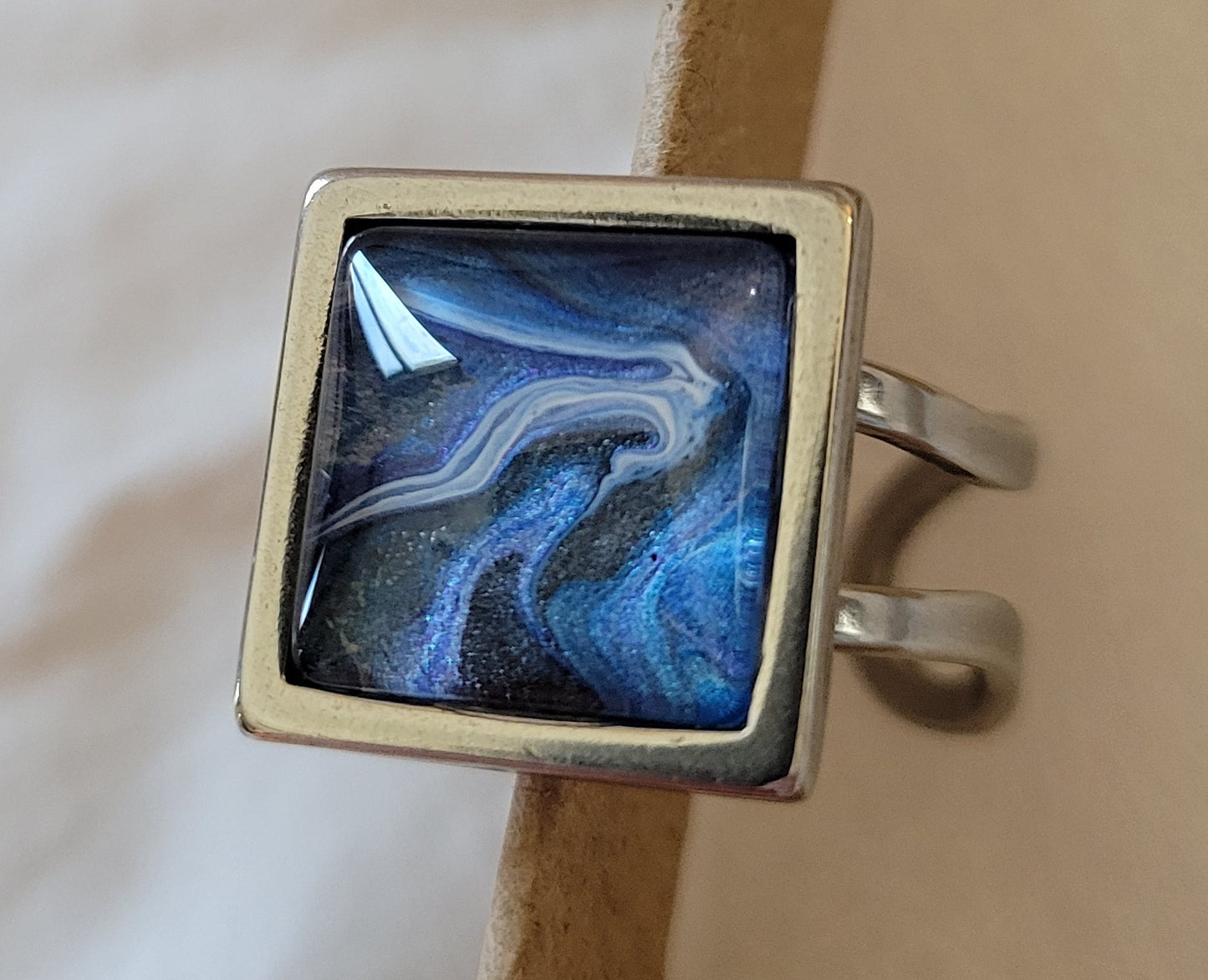 Square ring with looped bands rest against the edge of a cardboard jewelry box. The are are blues and silvers and purples and blacks that swirl together like a dark road stretching out into a cloudy ocean horizon.
