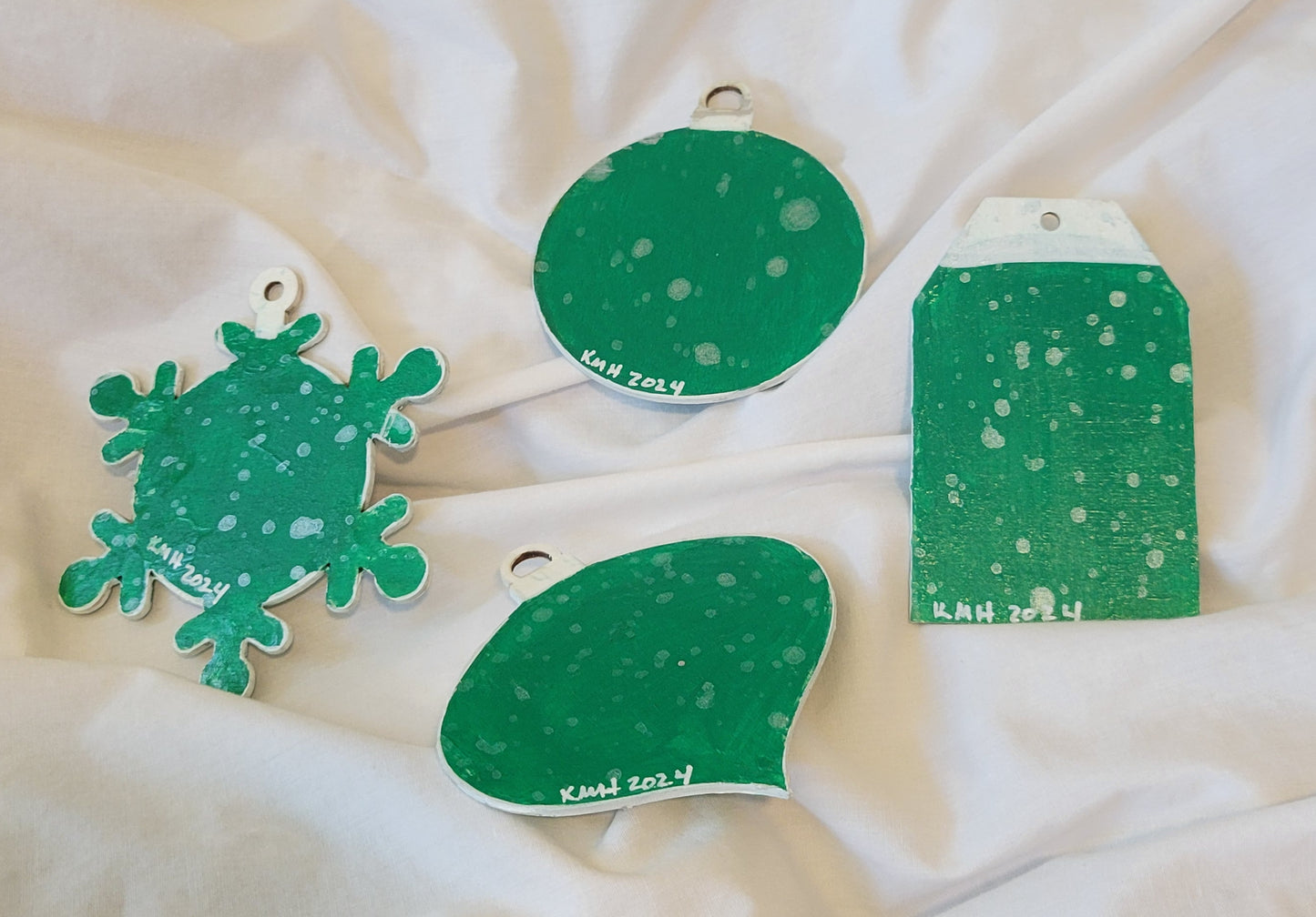 The reverse of the four wood-cut ornament shapes. They are all painted a bright green with white/pearl speckles. Towards the base of each ornament, "KMH 2024" is written in white.