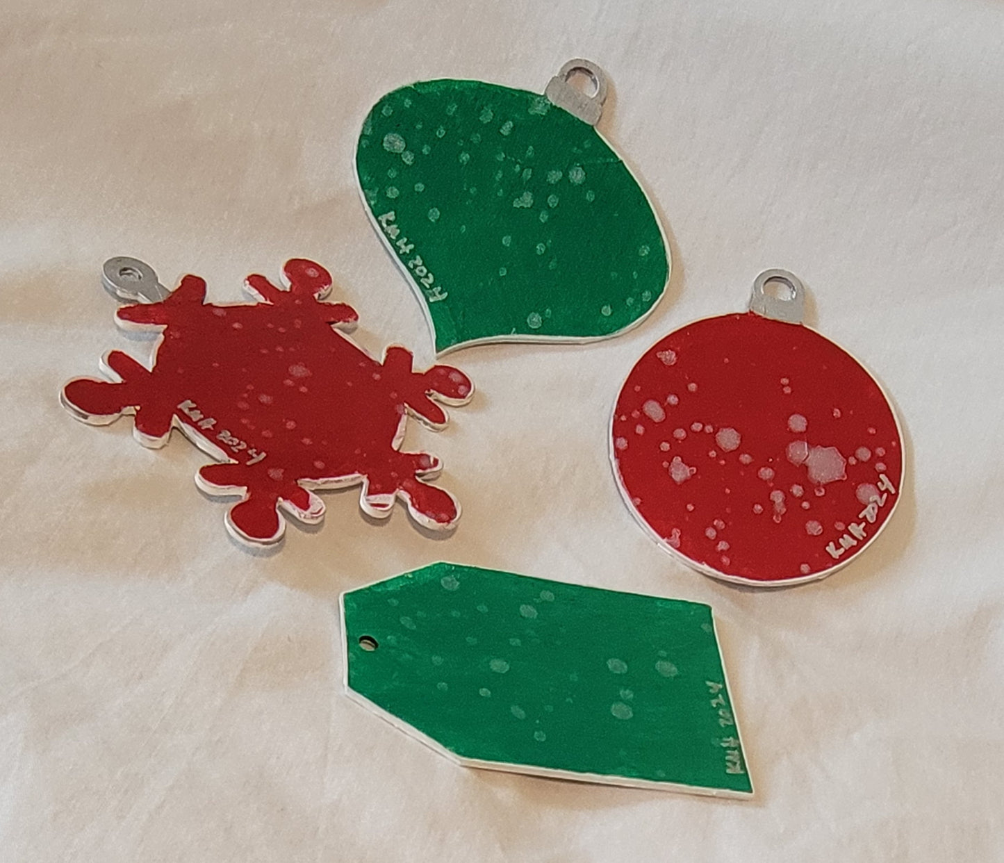 The reverse of the wood-cut ornaments. The snowflake and the ball are painted a bright red while the onion and the gift tag are painted a bright green. All feature white/pearl speckling with "KMH 2024" writen in white at the base of each.