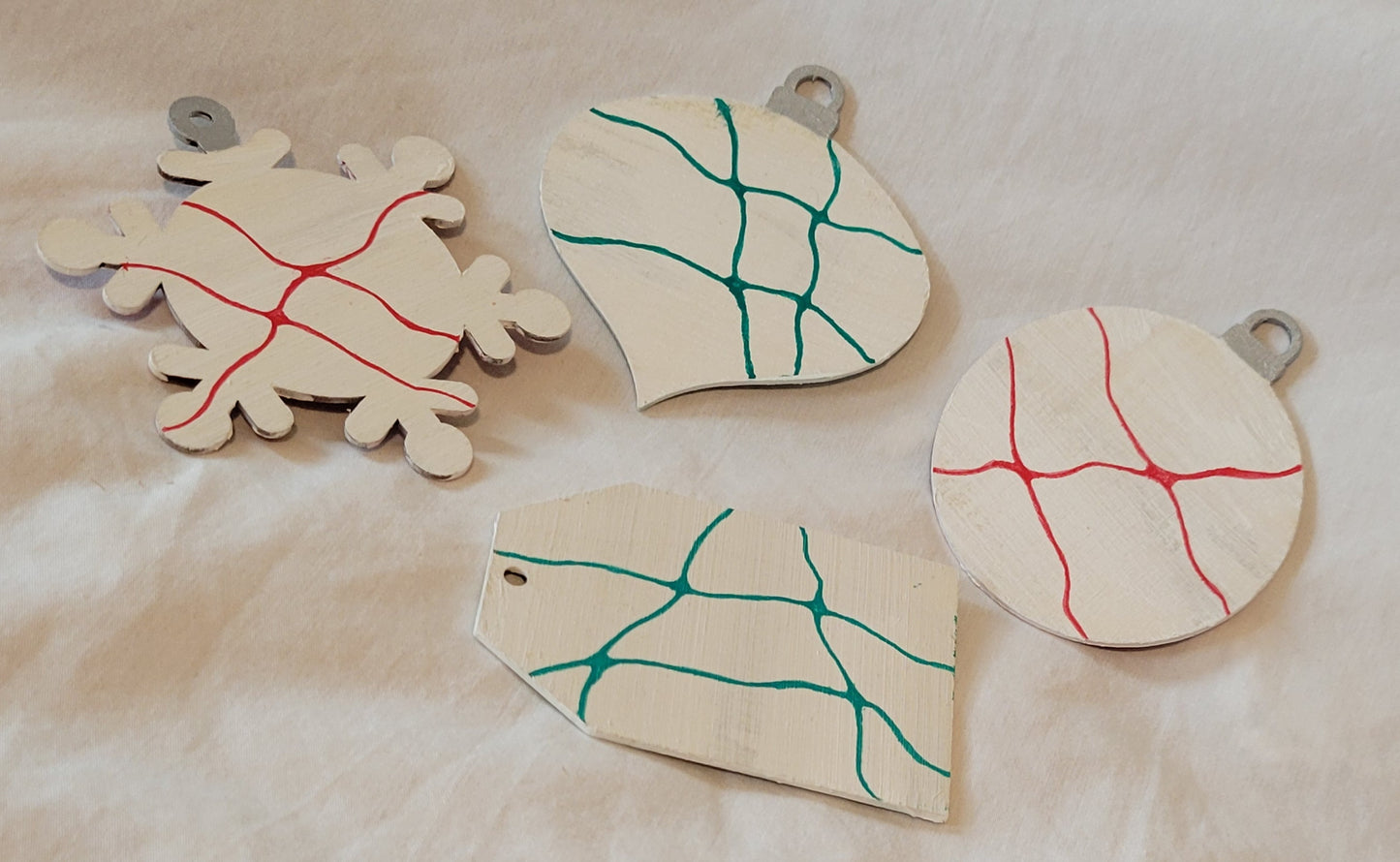 Four wood-cut ornaments in the shapes of a snowflake, oblong onion, ball, and gift tag rest on a white background. They are painted with a blend of white and silver. The snowflake and ball feature bright red neurographic lines. The onion and the gift tag feature forest green neurographic lines.