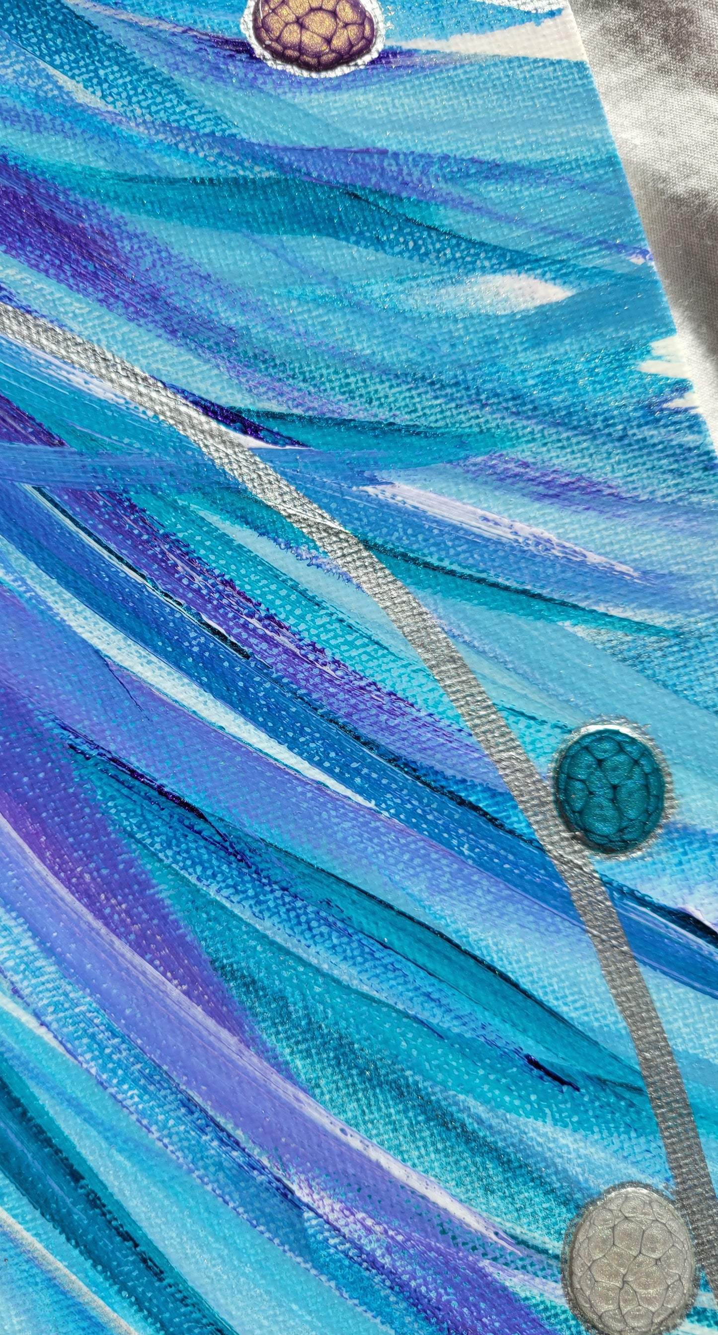 Closeup detail of the lines as they spill over the edge of the canvas, blues and teals and purples brushing and arcing over each other with a silver line crossing over them. Drops of purple, turquoise, and silver alkyd oil outlined in silver hug the silver line.