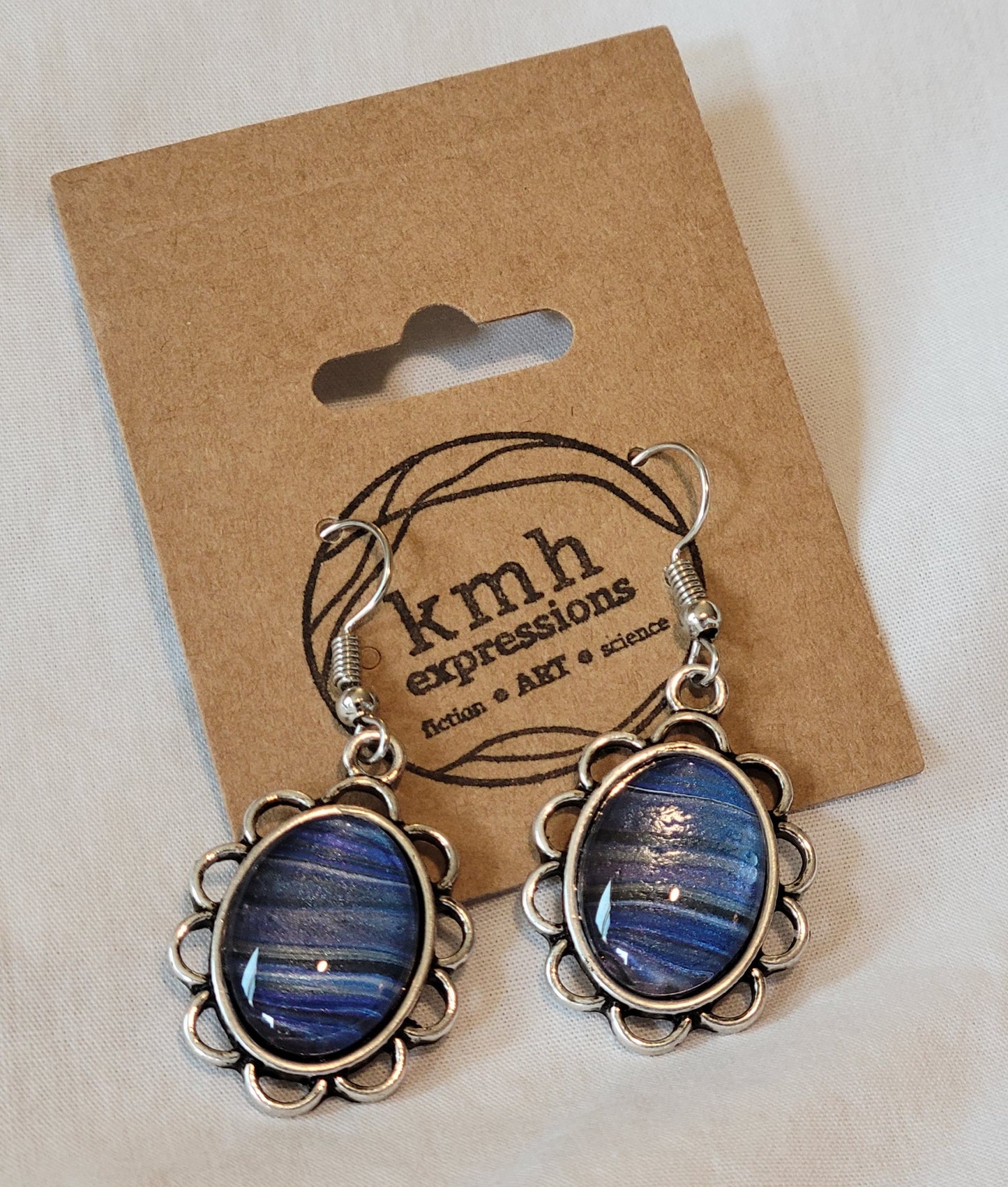 Scalloped silver filigree border a simple silver setting of these earrings dangling from a card stamped with the KMH Expressions logo. There are bands blues and purples with a few black and silver streaks.