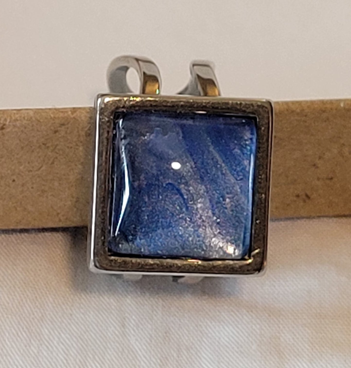 The ring features simple silver looped bands around the square setting for the cabochon. The coloring is predominantly deep blue with purple and silver highlights.