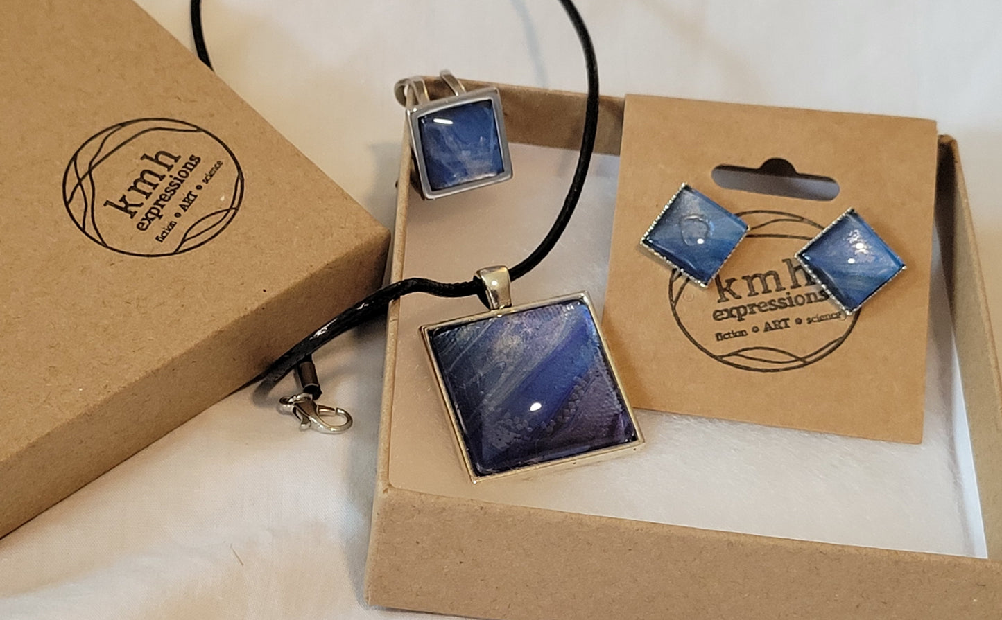 A jewelry set of necklace, ring, and earrings are displayed on white cotton padding in an open cardboard jewelry box. The set features blue, purple, and silver coloring that looks like more intense air currents.