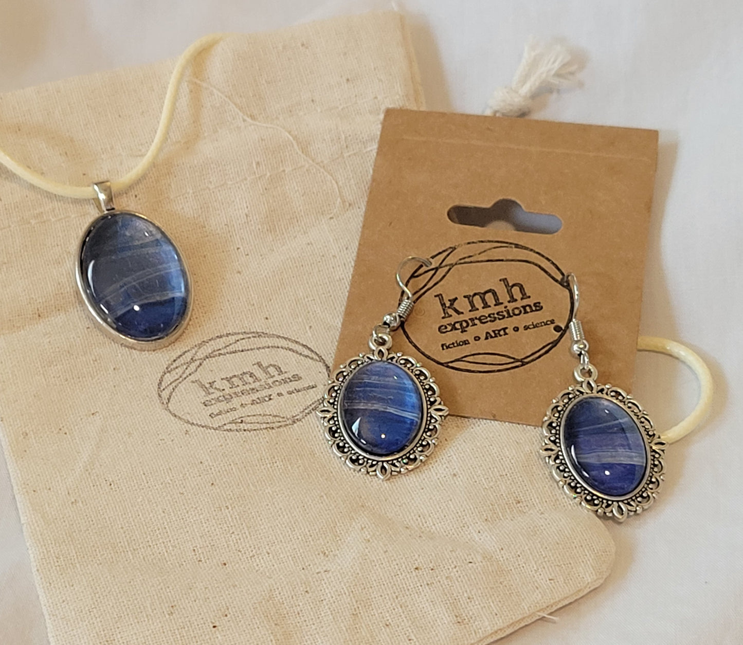 Bands of blues, silvers, and purples flow in harmony with each other across the jewelry. Oval necklace and earrings dangles on a backdrop of a muslin bag with my logo stamped on it. The earrings are dangling from a brown card with the kmh expressions logo stamped on it.