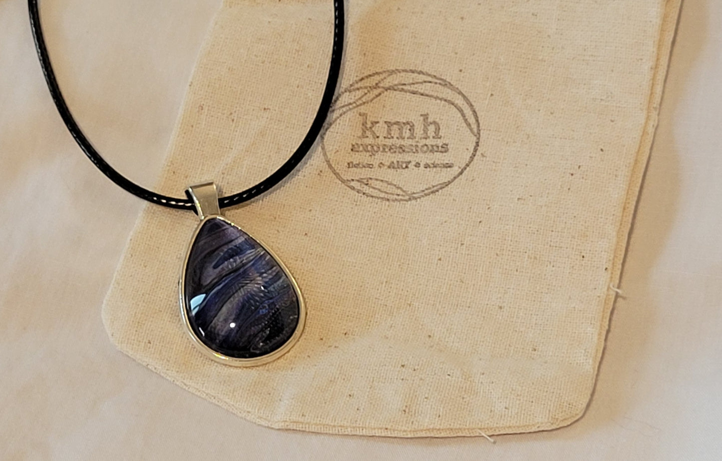 A teardrop pendant on a black imitation leather cord rests on a canvas jewelry bag stamped with the kmh expressions logo. The pendant is quite dark with purple deep blue and black, relieved with some silver banding.