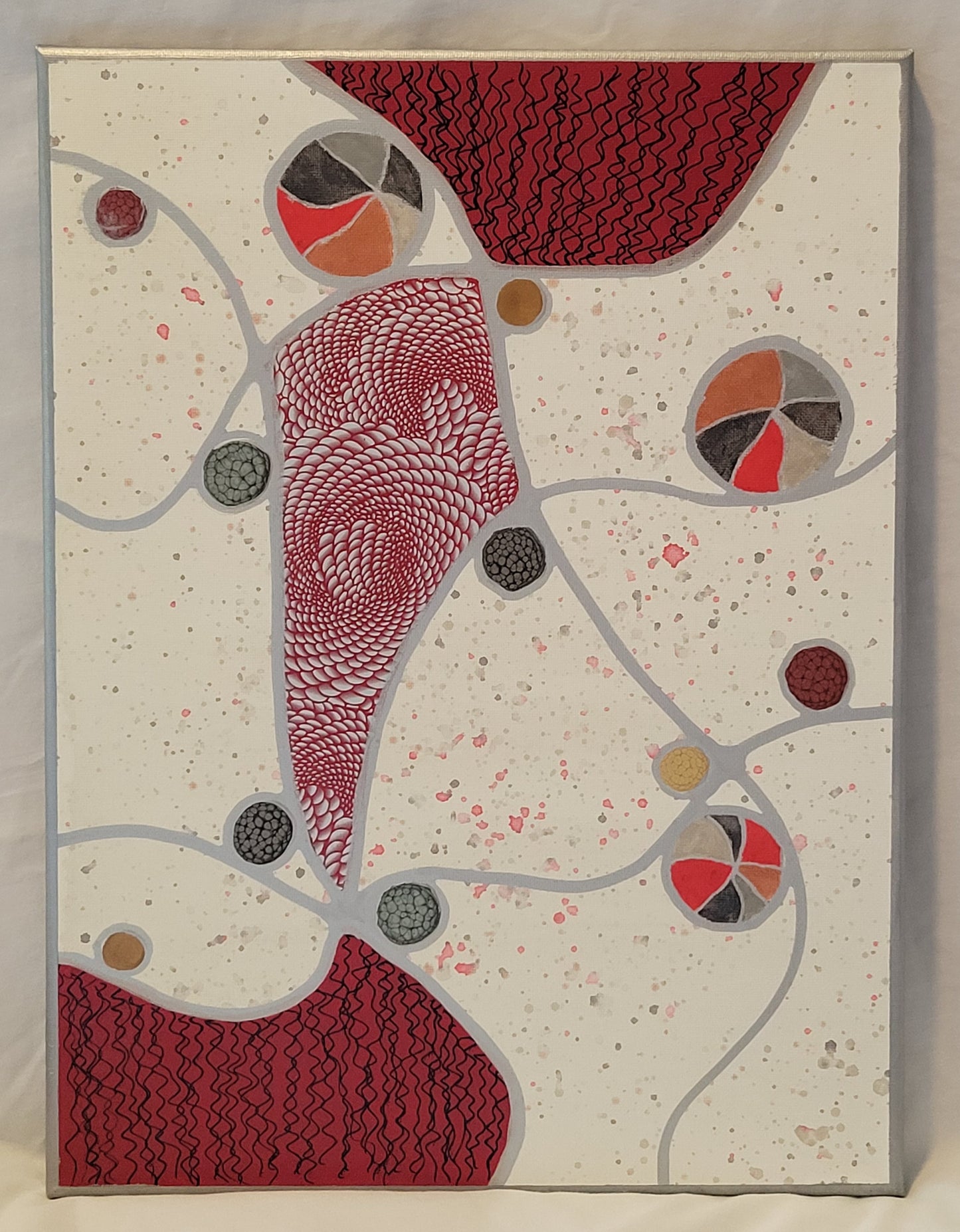 Silver lines arc and cross over a white canvas speckled with reds, grays, and silvers. Three of the shapes created by the intersecting lines are filled with red. Two of the three shapes at the top and bottom of the canvas have black squiggly lines. The central shape looks like a kite and is filled with a silver holographic foil in a psychedelic geometric pattern. Balls of red, black, silver, and gold alkyd oil or patterned acrylic tumble along the lines.