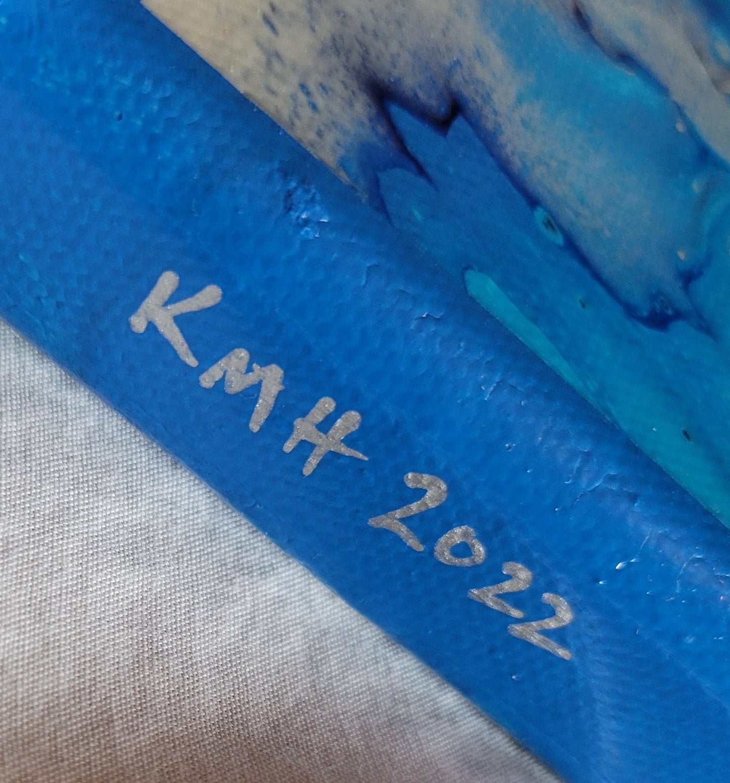 Closeup detail of the artist's initials and year of completion "KMH 2022" in silver against the deep sky blue on an edge of the canvas. Some of the browns and blues of the composition can be seen.