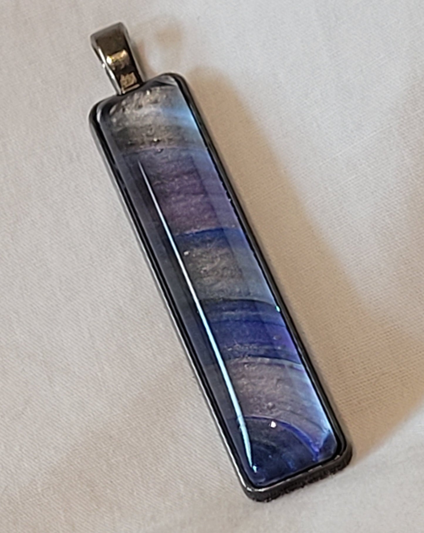 Rectangular pendant in a dark gray stainless steel. The bands of silver, space gray, dark blue, and purple, seem to arc around the lower left corner of the pendant like the rings of a planet.