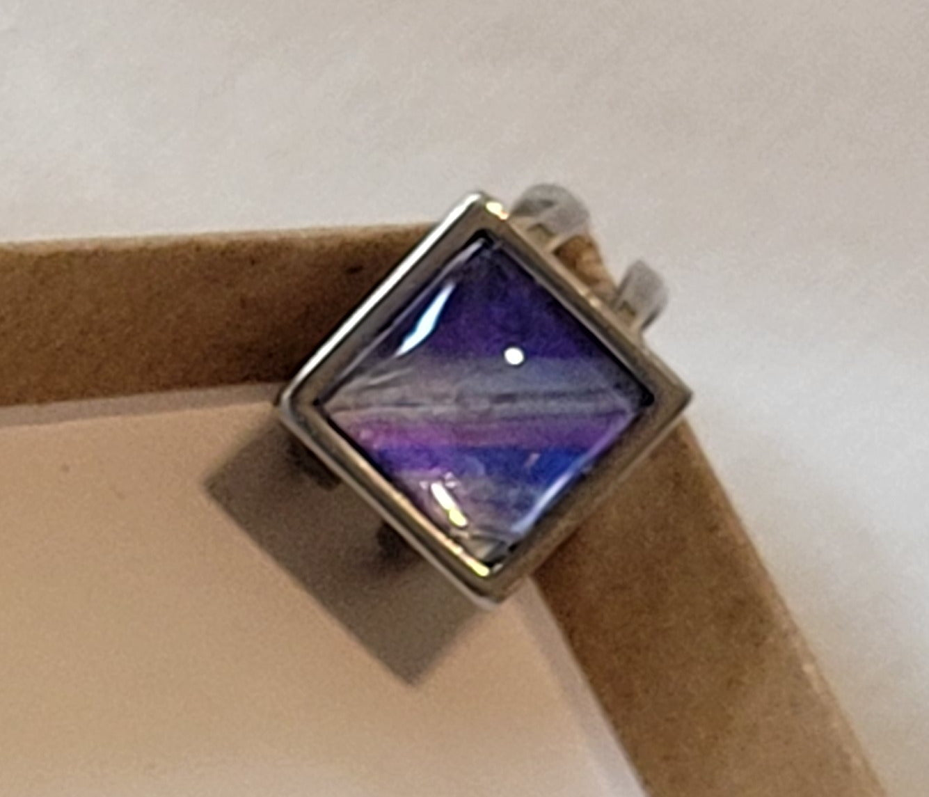 Looped bands of a simple stainless steel square ring resting on the edge of a cardboard box. There bands of dark purple, silver, lighter purple, and dark blue are threaded through with thin lines of space gray.