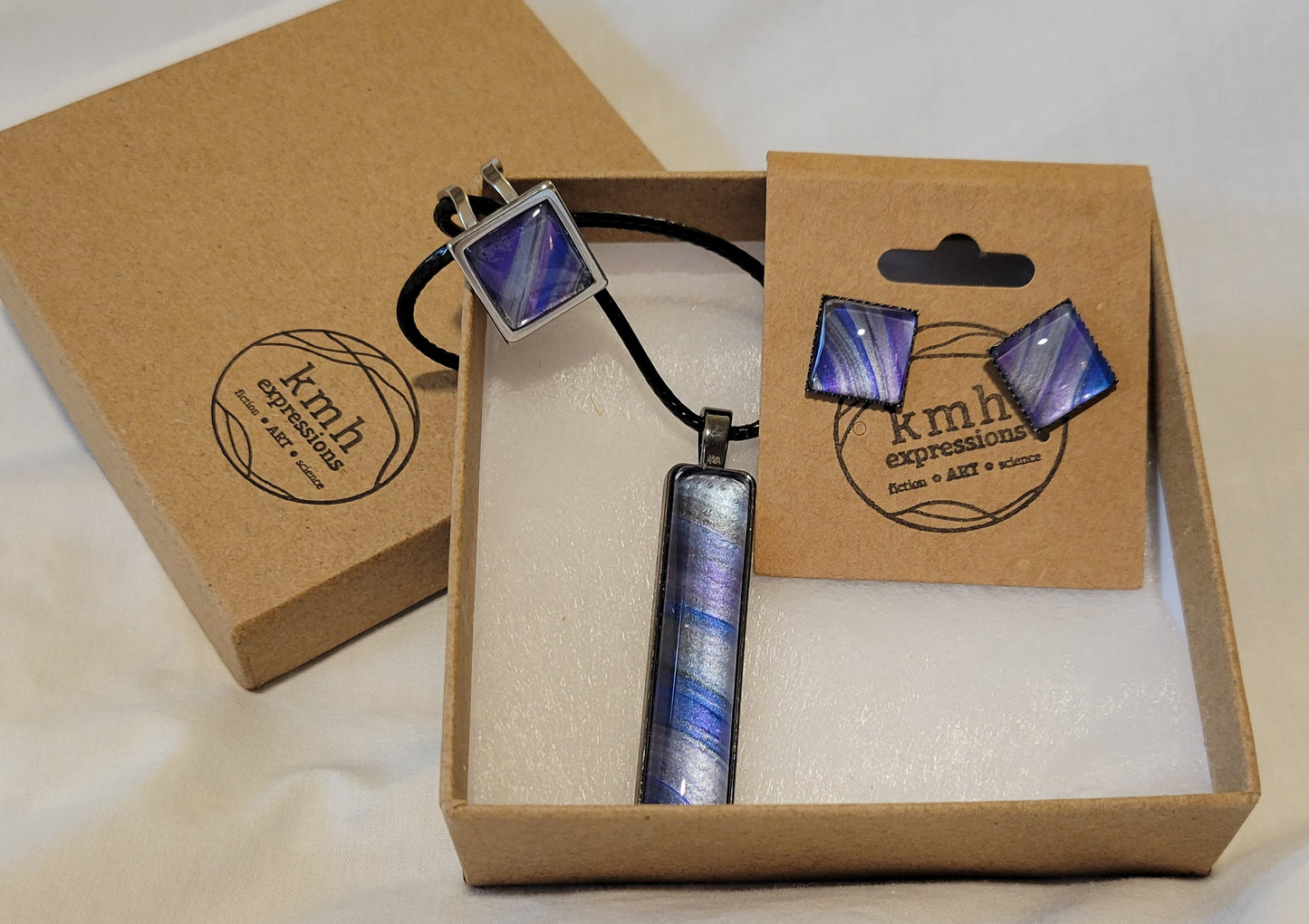 A three-piece set of jewelry (necklace, earrings, and a ring) resting in a cardboard box and earring card stamped with my kmh expressions logo. The pieces feature bands of silver against dark blue, space gray, and purple.