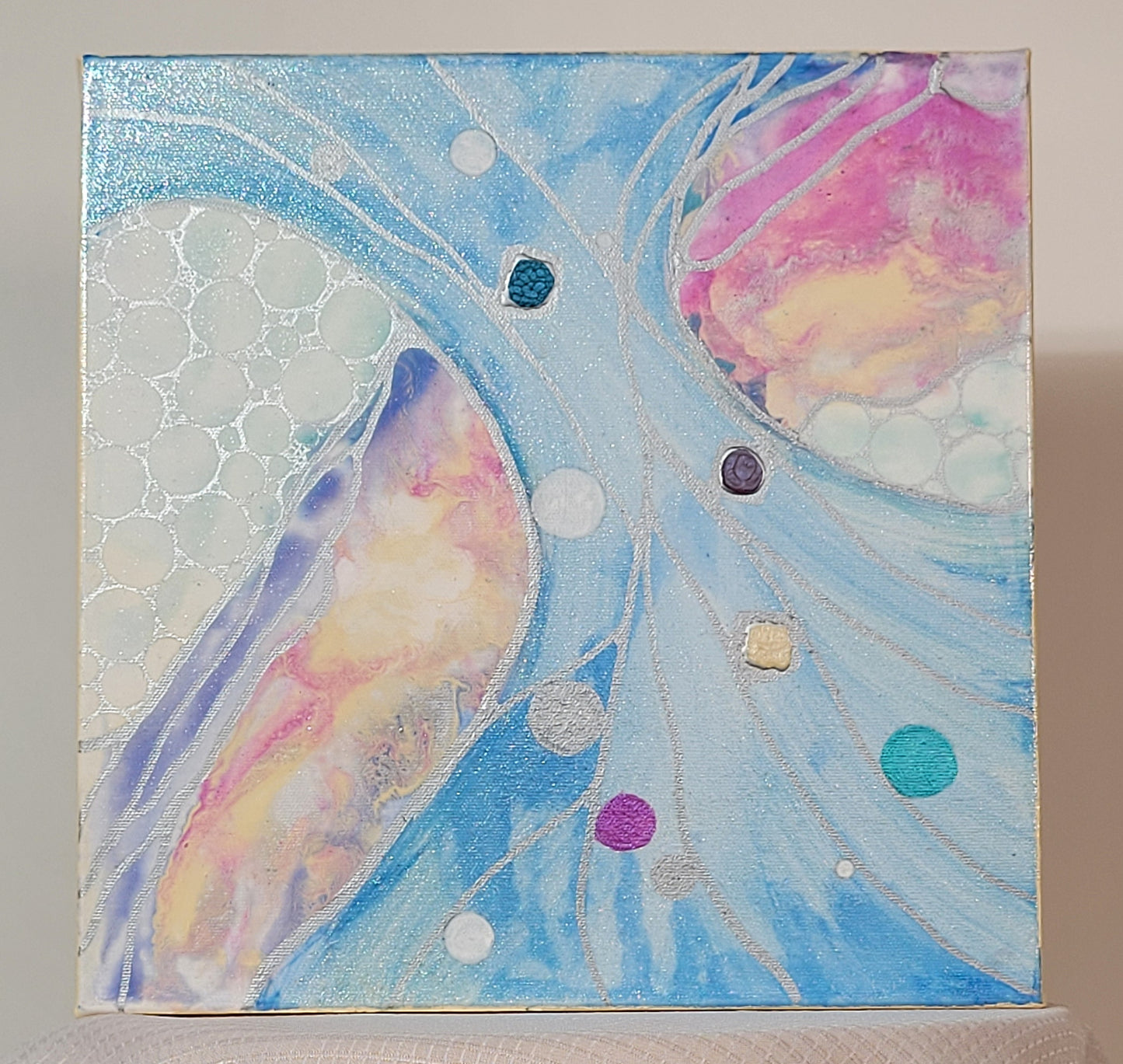An hour-glass shape of distressed sky blue stretches across a diagonal of the canvas. Underneath it, an arc of fluid acrylic stretches along the other diagonal in violet, pink, and peach. Silver circles are crowded together over a distressed white and turquoise background in the spaces not otherwise covered by fluid acrylic or the hourglass. Silver lines thread through the hourglass and at the edges of the fluid acrylic. Alkyd oil and acrylic circles in turquoise, silver, white, and purple dot the hourglass