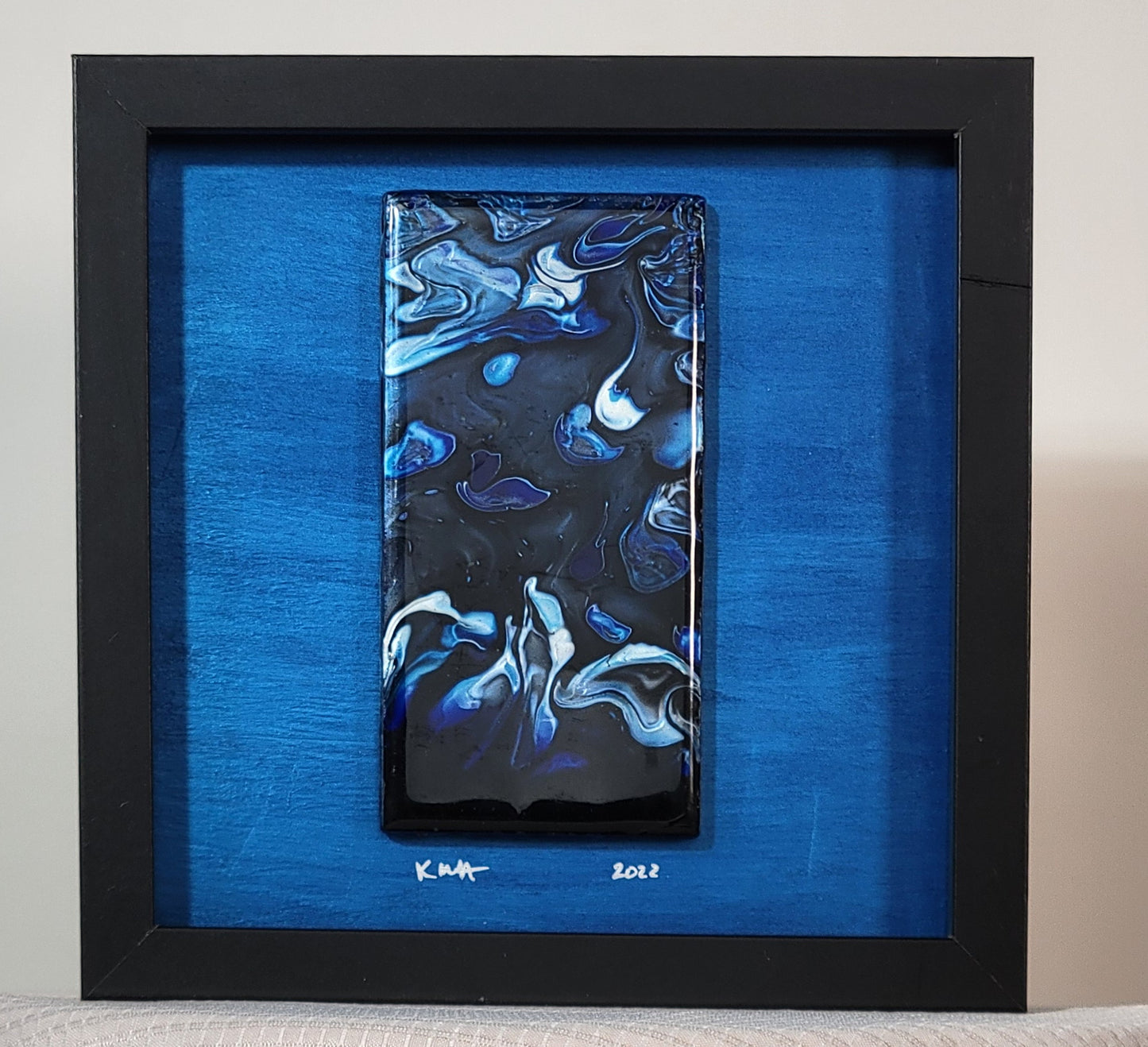 Shadow box with a black frame and a royal blue background with a tile mounted on it; just under the tile, the artist has signed their initials KMH and the date 2022 in silver. The tile has a black background with flowing, distorted splotches of silver, light blue, and dark blue that seem to float and flow and disturb each other.