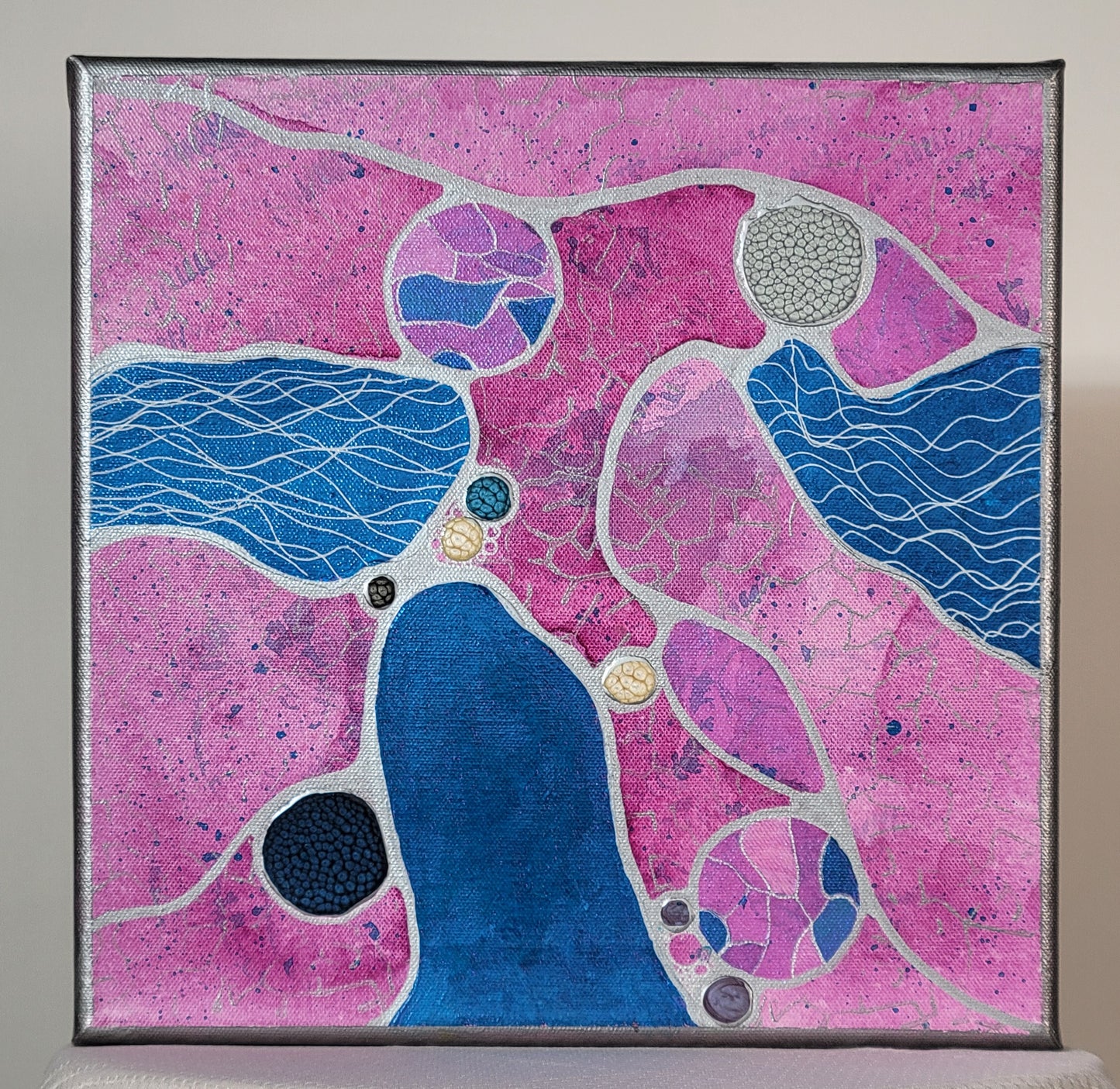 Shiny blue amoeba-like shapes and bubbly gray, white, black, purple, and dark blue circular spore shapes ooze out of two cracked and blue-stained abstract petri dishes. Everything is spilled onto a background of a pink-hued nutrient bath effect, with a hint of blue speckles and patterned silver tread. Heavy silver lines like cilia connect everything in a neurographic style. Two of the amoeba shapes have white cilia within them while the third has a hint of holographic pebbling.