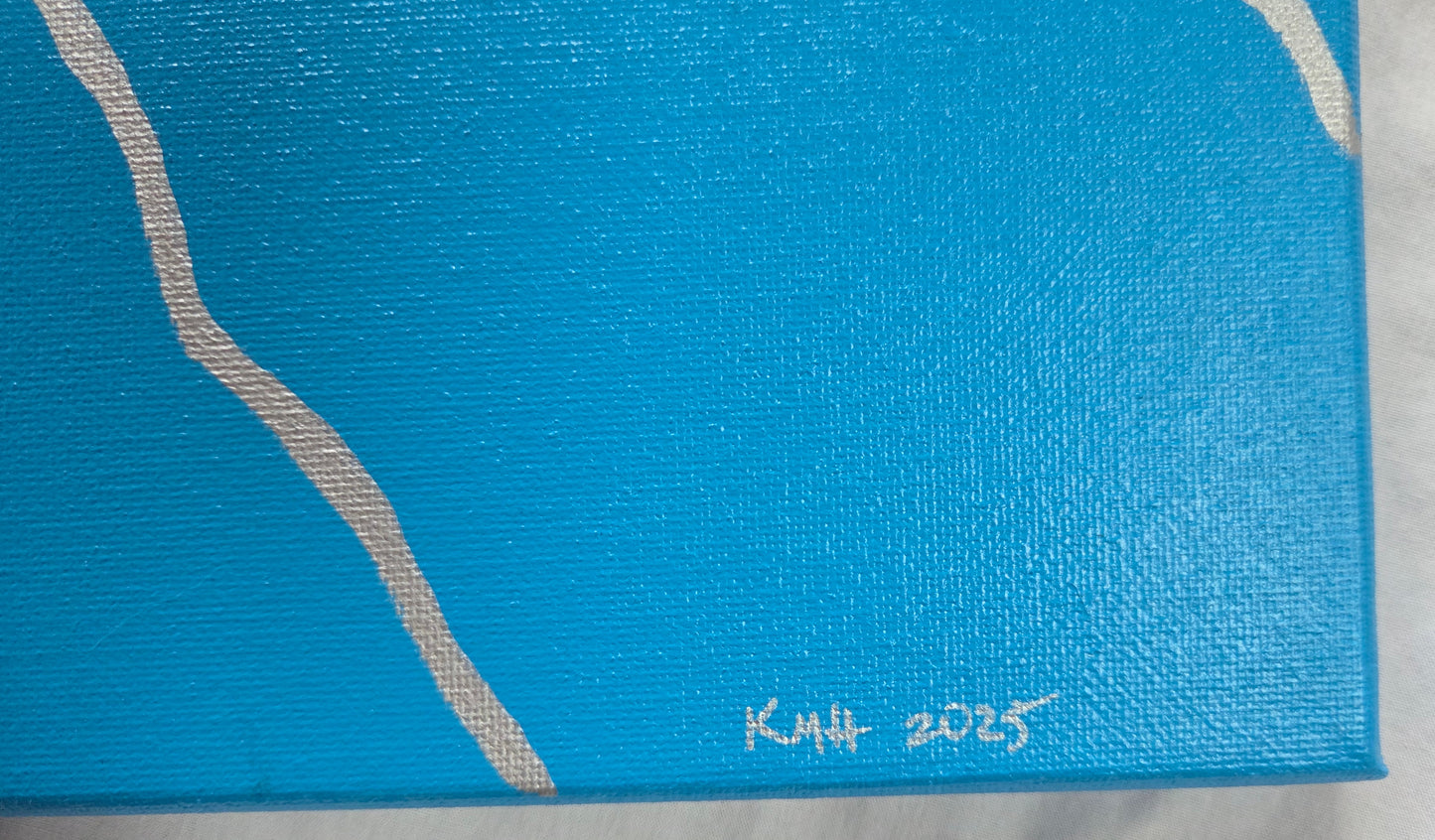 Close up detail of the bottom-right corner of the trans pride flag showing thick silver lines and the artist's initials and year of completion in silver.