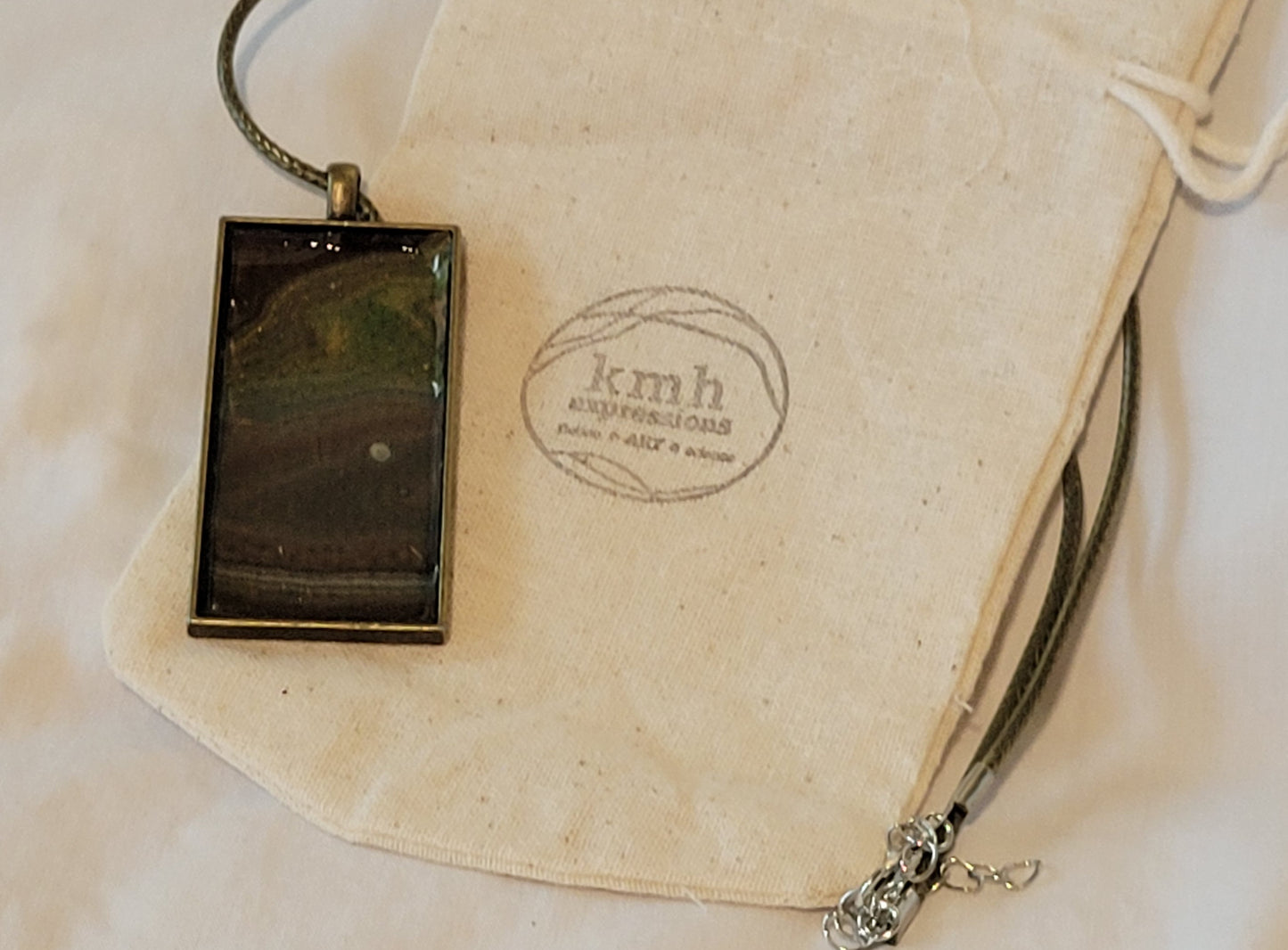A rectangular pendant with a bronze alloy setting and a moss green imitation leather cord resting on a muslin bag stamped with the kmh expressions logo. The pendant has a variety of earth tones in brown, gray, green, and ochre stacked on each other that call to mind the banding patterns of the Grand Canyon. The pendant backing has dotted depressions that come through the acrylic, looking like rain.