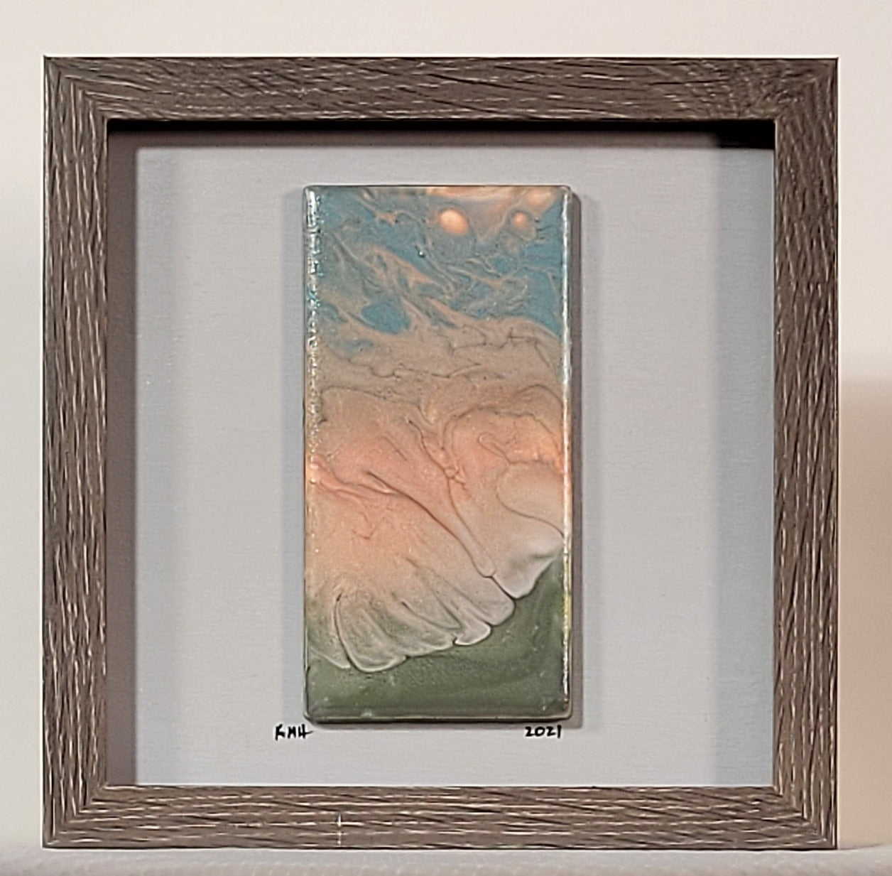 Shadow box with a brown wood grain frame and an off-white background with a tile mounted on it; just under the tile, the artist has signed their initials KMH and the date 2021. On the tile: Aqua blue, sandy brown and bronze-red all flow and twist around each other. The bronze-red also swirls with a chalky white to intrude upon a moss green.