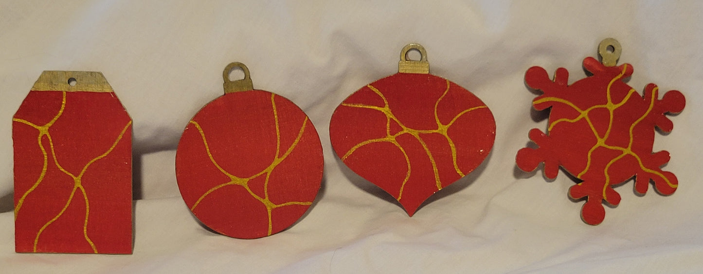 Four wood-cut ornaments in the shapes of a gift tag, ball, oblong onion, and snowflake rest on a white background. They are painted with a blend of bright red and gold. They feature thin gold neurographic lines.
