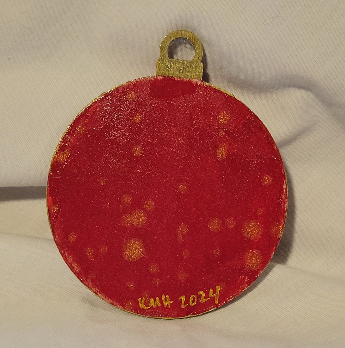 The reverse of the ball woodcut ornament. It is painted bright red and speckled with bright gold. At the base of the ornament "KMH 2024" is written in thin, bright gold.