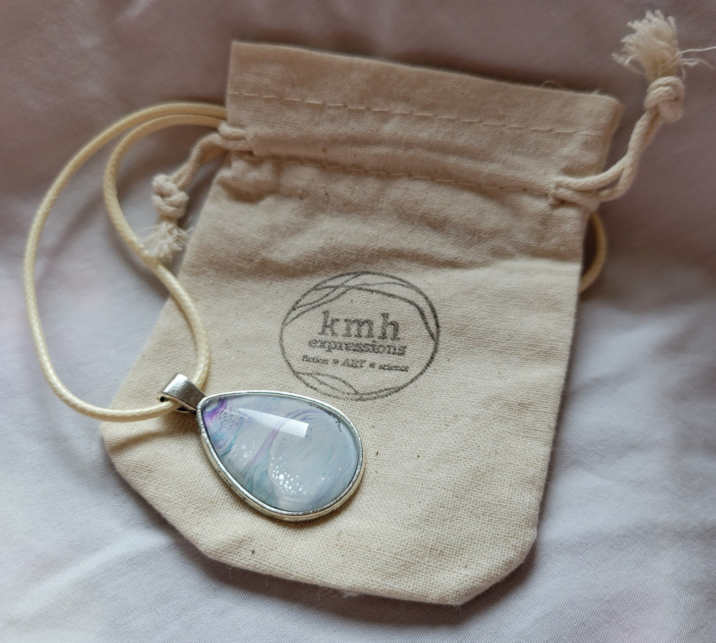 A simple stainless steel oval pendant with a cream-colored imitation leather cord rests against a muslin bag stamped with the kmh expressions logo. The pendant as an overall white tone with strong threads of a light green-blue and purple.