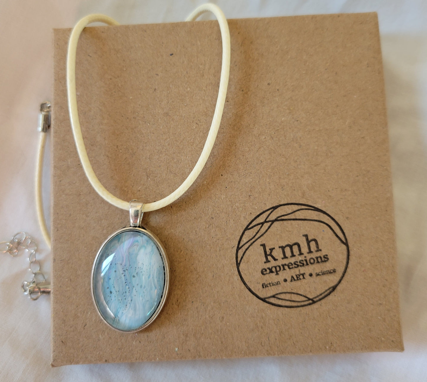 A simple oval stainless steel pendant with a cream-colored imitation leather cord rests on a brown cardboard jewelry box stamped with the kmh expressions logo. The pendant overall has a light teal color threaded with white and slight hints of purple.