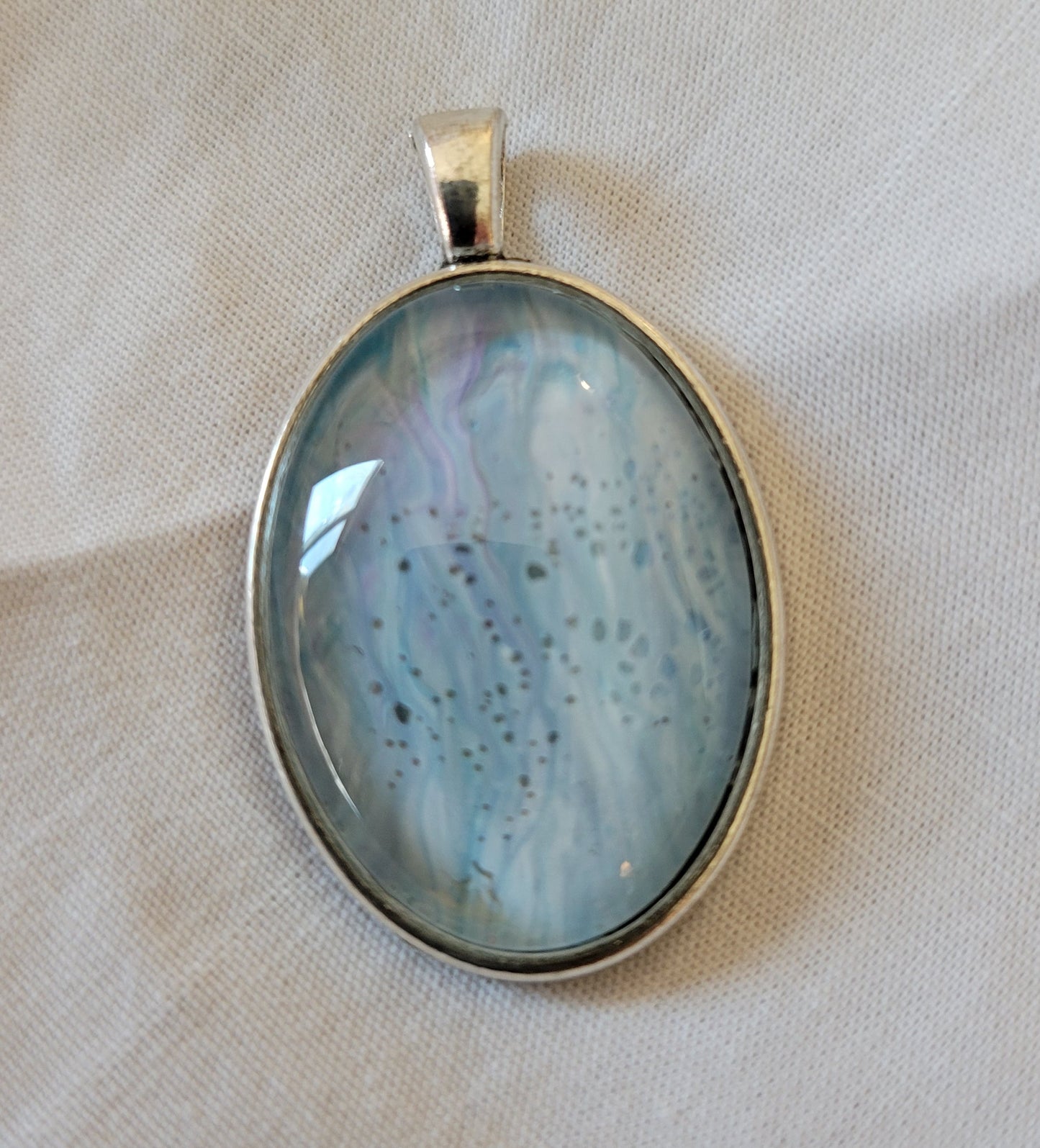 A simple stainless steel oval pendant rests on a white background. There are large streaks of light teal threaded with purple and clouded with white.