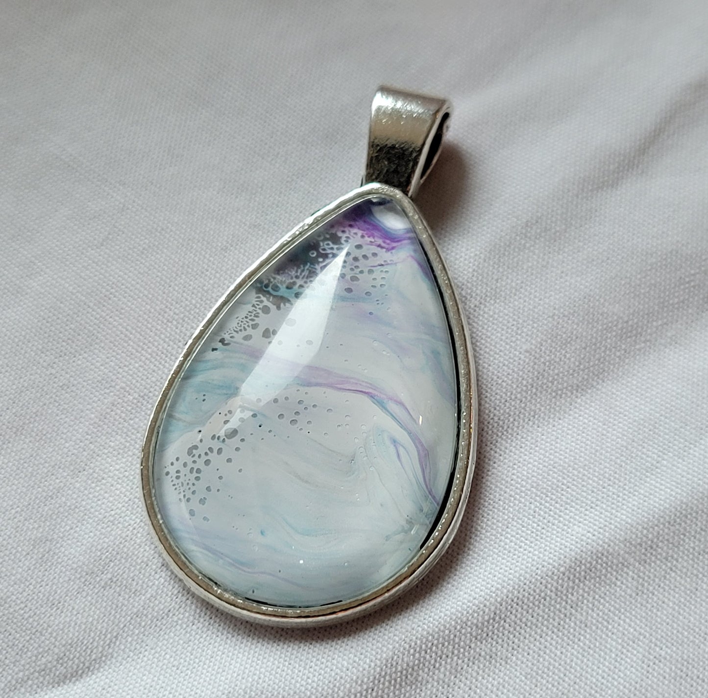 A simple stainless steel oval pendant rests on a background of white. The cabochon has a white tone that looks like foamy ocean spray with hints of green-blue waters underneath and dark purple threads into the distance that look like a stormy sky.