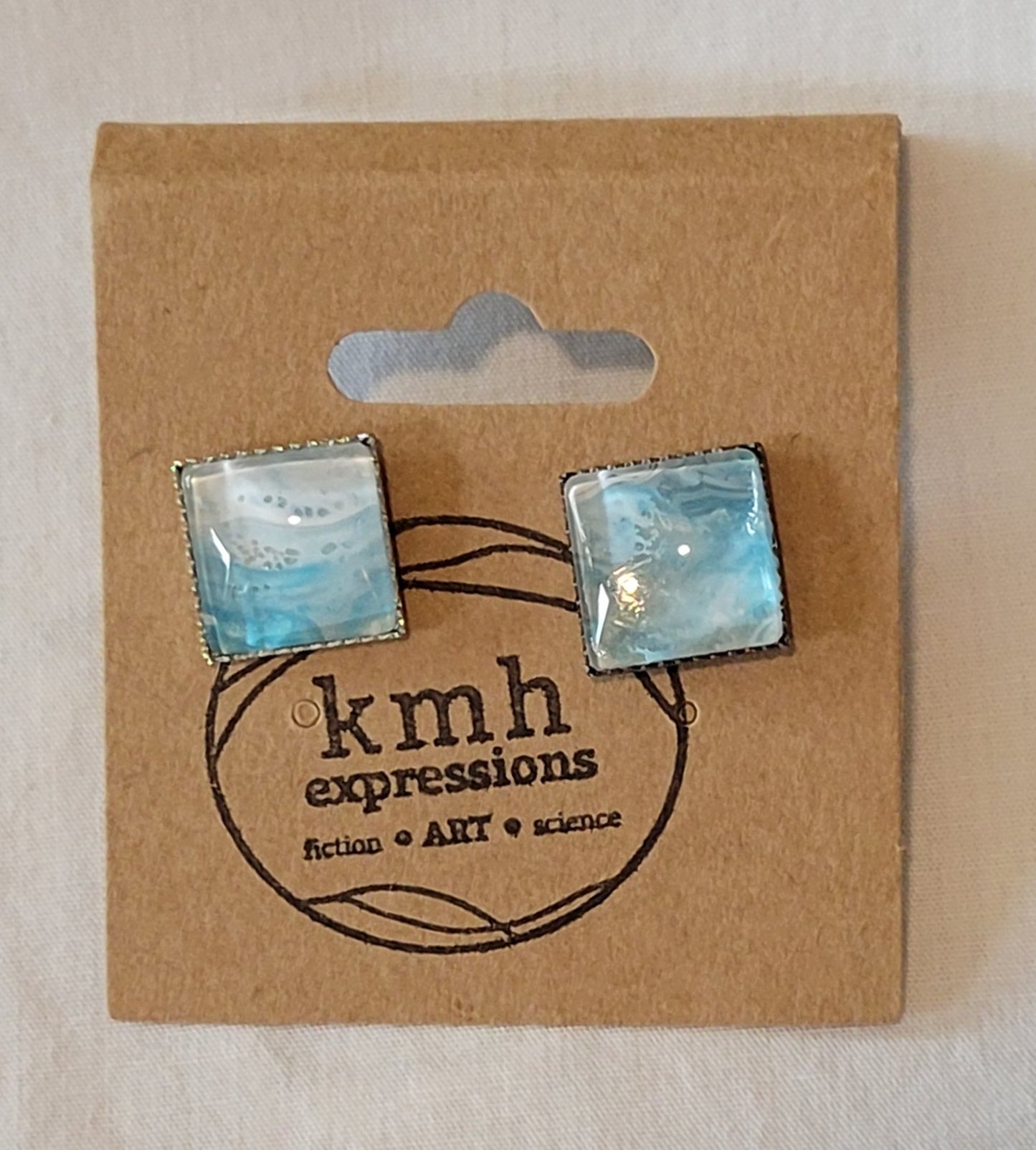 Simple black pierced studs on a brown card stamped with the kmh expressions logo. The cabochons are a blend of white and teal like waves in the shallow ocean surf.