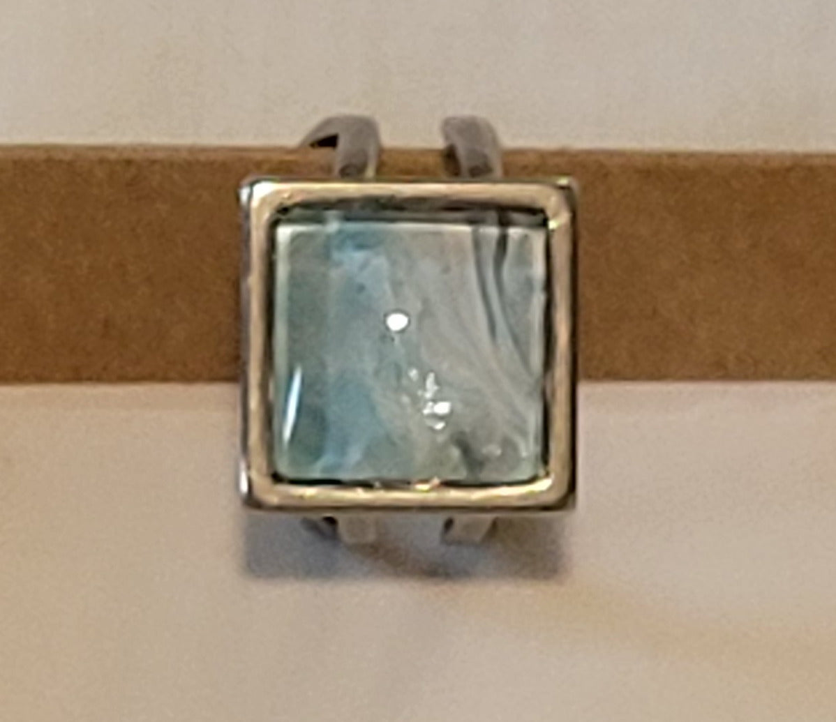 A simple stainless steel square ring with looped bands rests on the end of a cardboard jewelry box. The cabochon features white waves over a teal background.