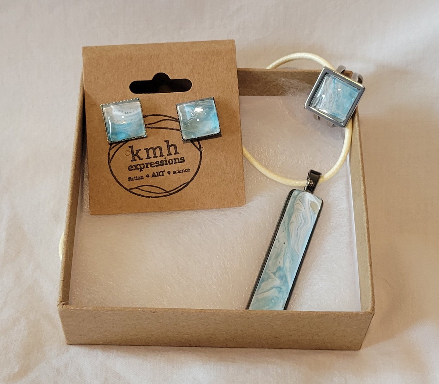 A jewelry set including earrings, ring, and necklace rest on gauze in a brown cardboard jewelry box. The colors of the set are predominantly seafoam green and teal threaded through with white.