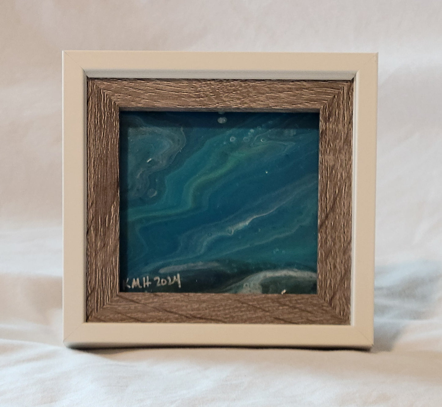 A picture frame with white outer framing and brown wood-grain inner framing. The featured picture is a fluid art square featuring shades of blue, light green, and white banded in a rippling wave pattern. In silver in the lower left corner are the artist's initials "KMH" and the year completed "2024."