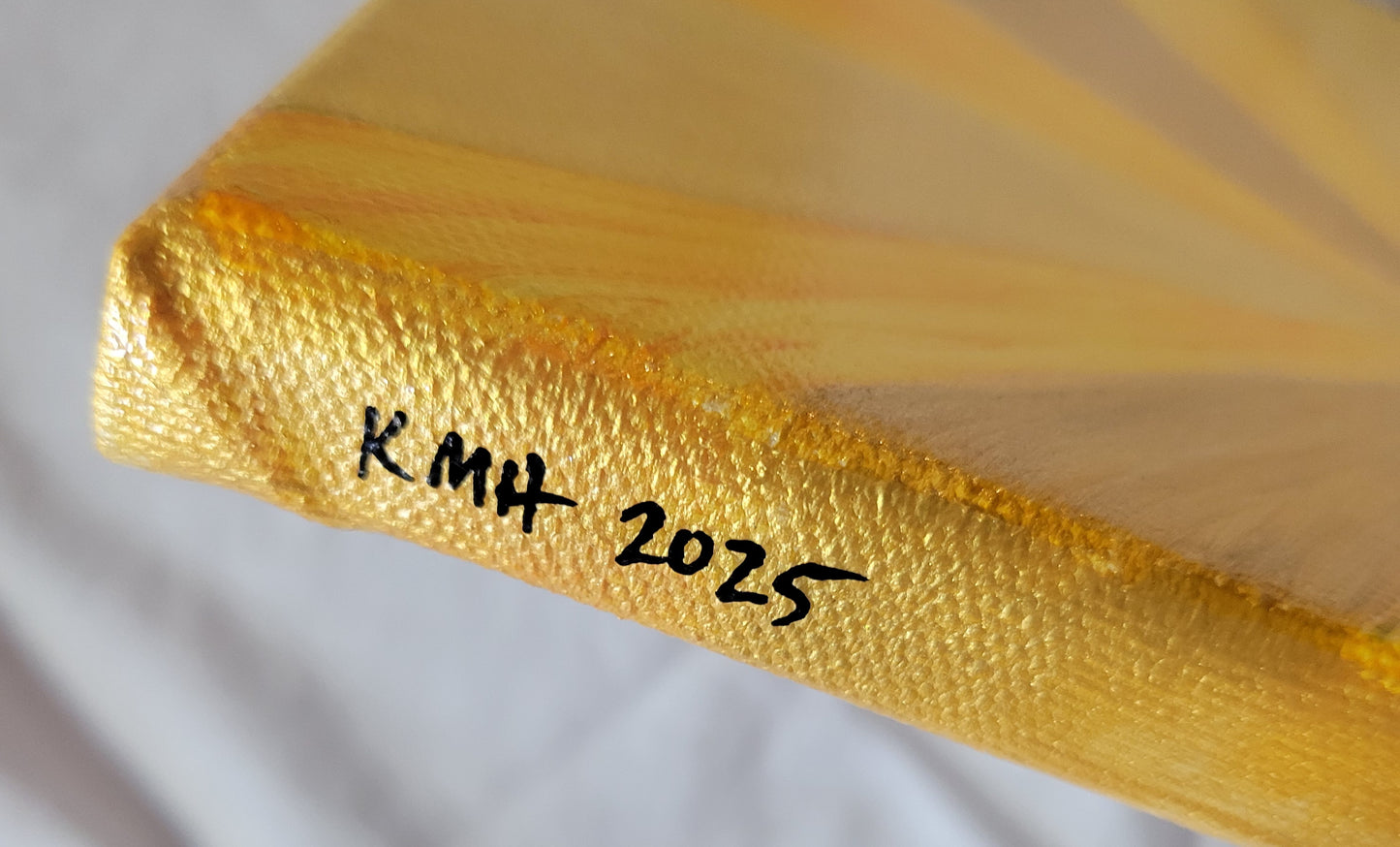 Side angle view of the golden edge of the painting with the artist's initials and year of completion "KMH 2025" written in black.