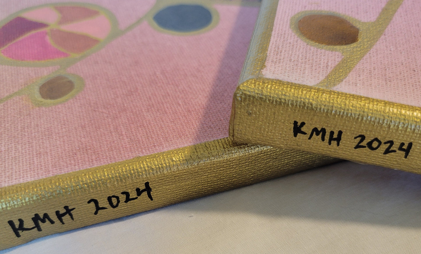 One canvas resting at an angle against another. Both are viewed from the gold-painted edges where "KMH 2024" in black can be seen. The distressed pink and gold backgrounds of the canvases can be seen along with multicolored balls and alkyd oil spheres.