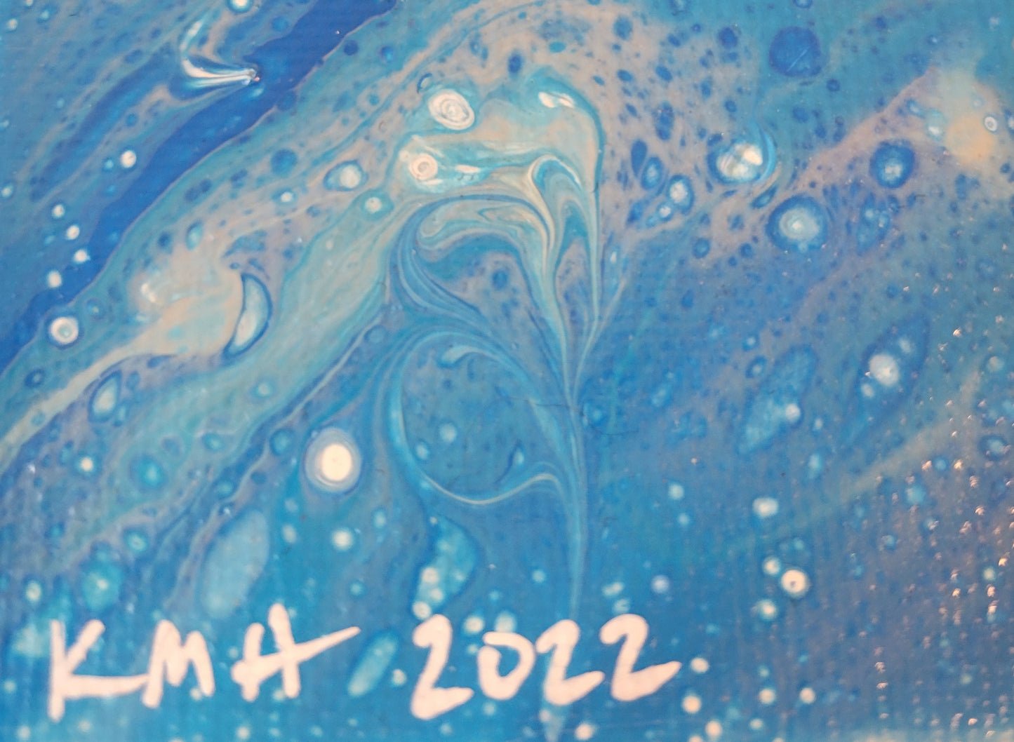 Close-up detail of the base of the canvas with "KMH 2022" written in white against the background of banding, bubbling, and swooping blues and whites against a sandy brown base.