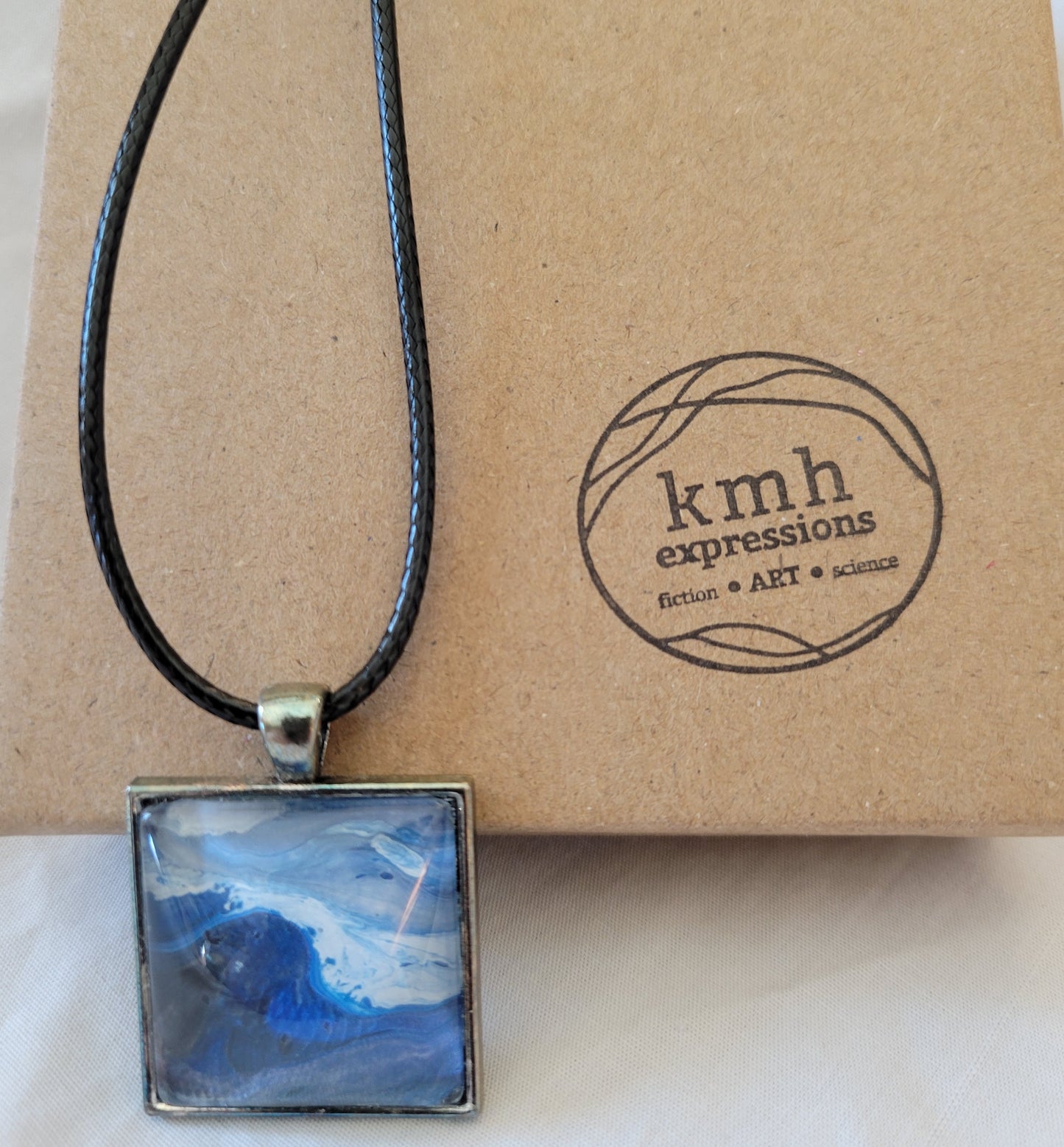 A simple square stainless steel pendant with a black imitation leather cord rests against a brown cardboard jewelry box stamped with the kmh expressions logo The cabochon features a deep blue cresting, frothing wave against a gray blue white stormy sky.