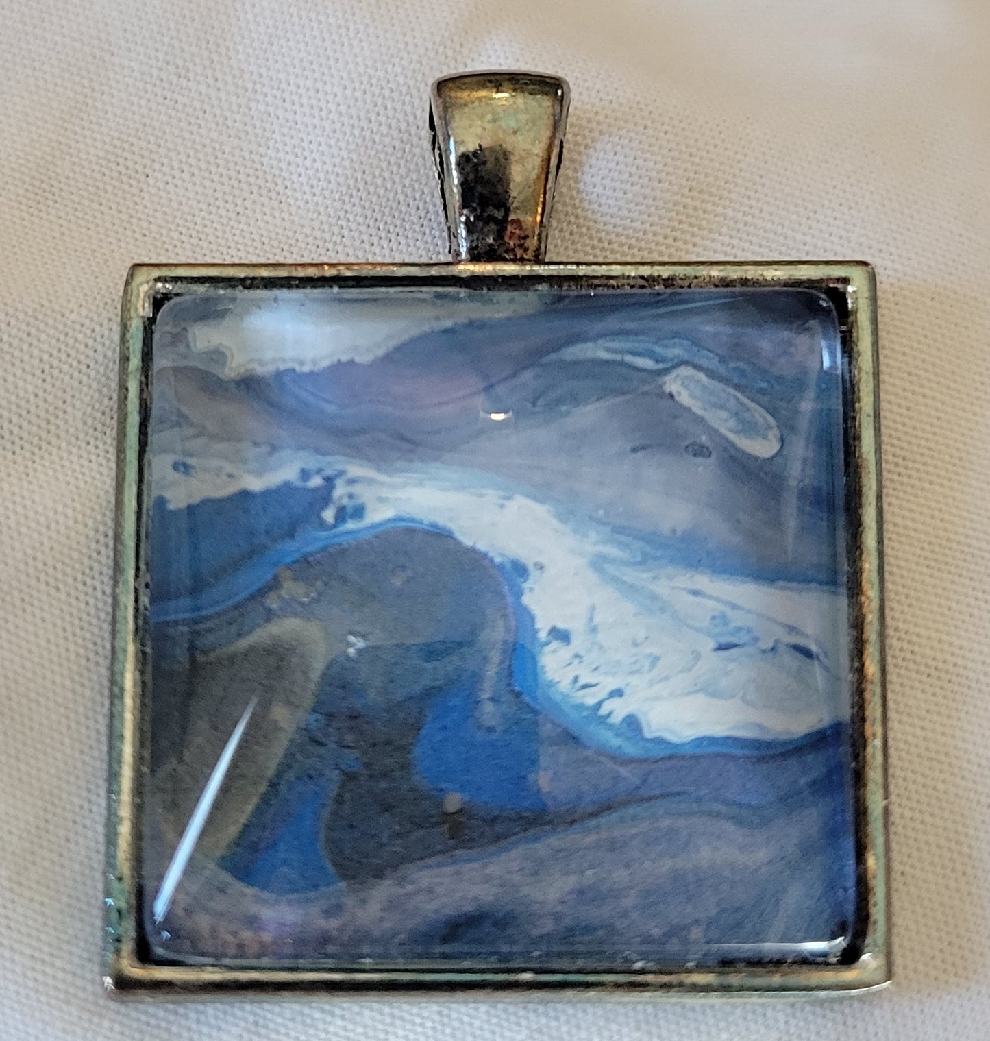A simple square stainless steel pendant rests on a white background. The arcing blues with dark undertones is more clearly seen. White at the end of the dark blue crests bubbles slight to give an ocean spray look. The dark purple, blue, gray and white of the rest of the background looks like thready, stormy clouds.