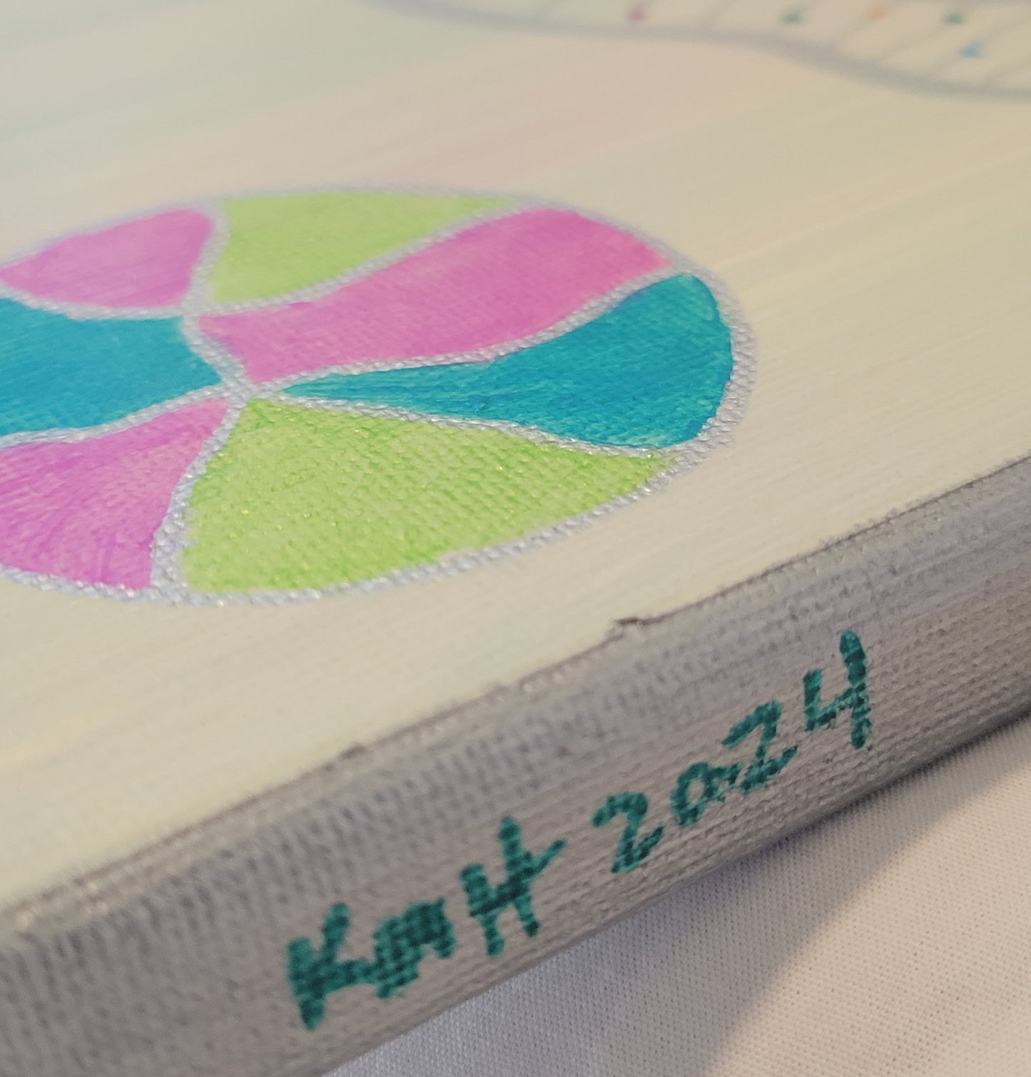 Side-angle shot of the canvas, showing the green, blue, pink, and silver circle and a silver painted edge with "KMH 2024" written in teal.