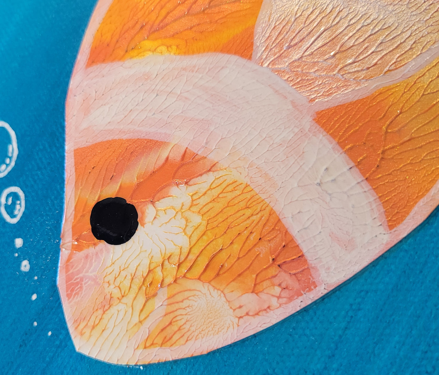 Close-up of the clownfish, showing the scaly blend of orange, gold, and pearl with clownfish pattern painting in white and pearl with a black alkyd oil dot for an eye. There are drawn white bubbles floating from the fish mouth.