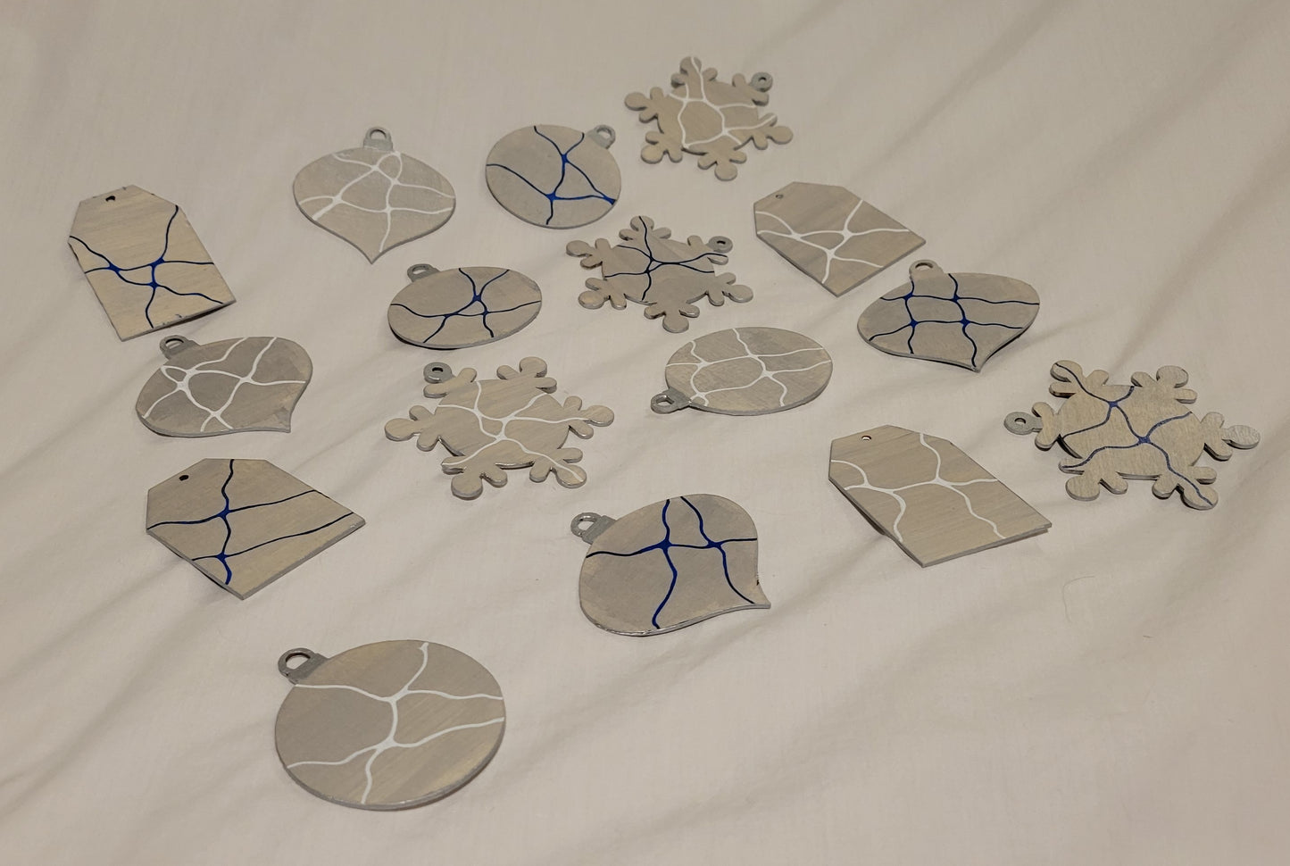 All 16 (4 sets of 4) ornaments rest on a white background. They are all painted with a blend of silver and white and all feature a unique detail of line work in either white or royal blue.